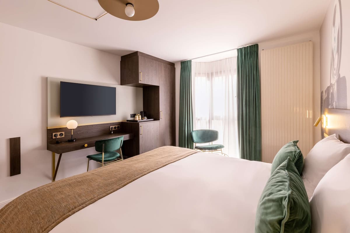 A hotel room with a bed, a desk with a chair, a TV mounted on the wall, and a window with curtains.
