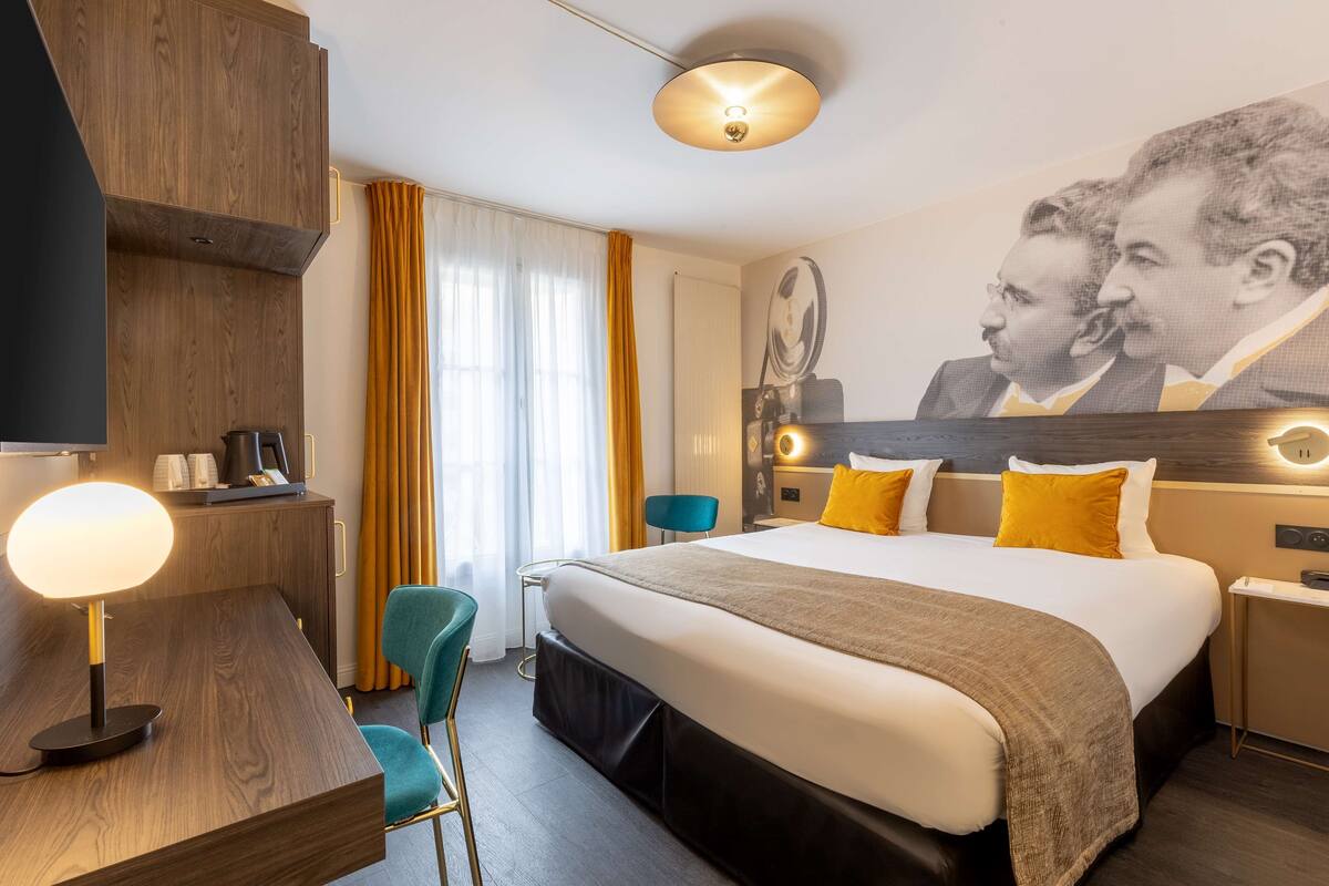 A hotel room with a large bed, a desk with a lamp, a chair, a wardrobe, and a wall-mounted TV.