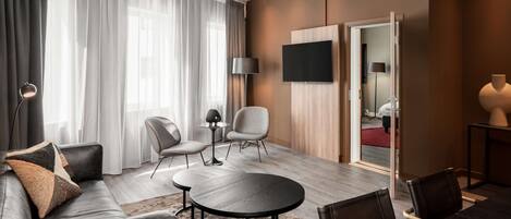 A modern hotel room with a sofa, two armchairs, a round table, a TV, and a large mirror.