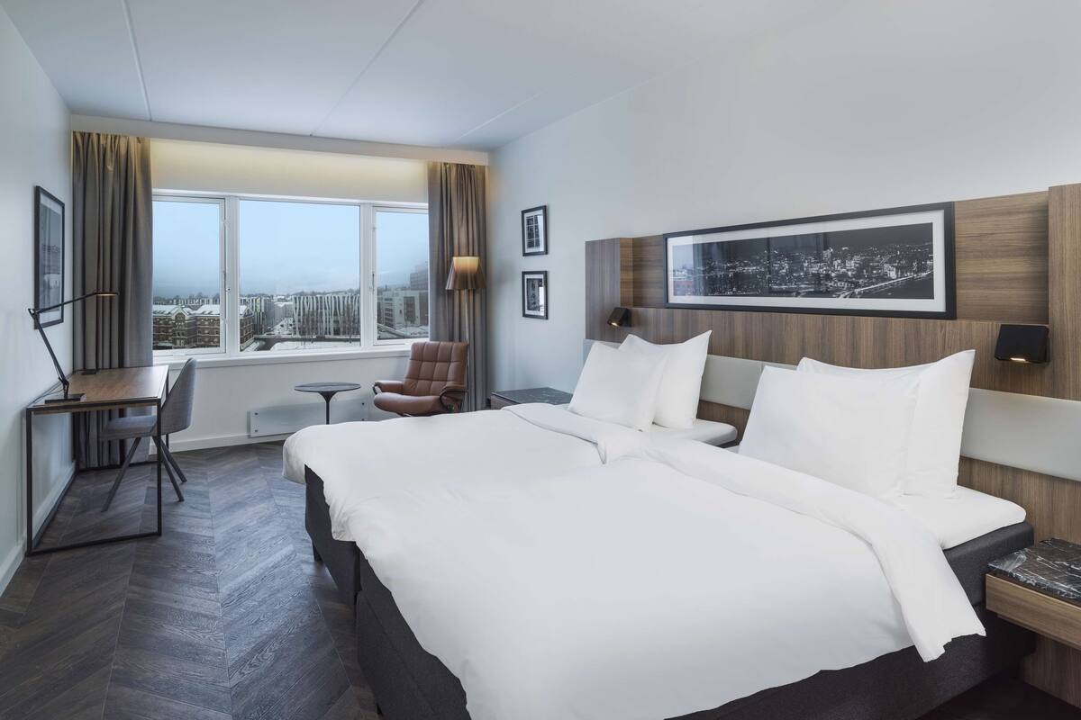 Quarto de hotel com uma cama grande, uma mesa, uma cadeira e vista para a cidade pela janela.