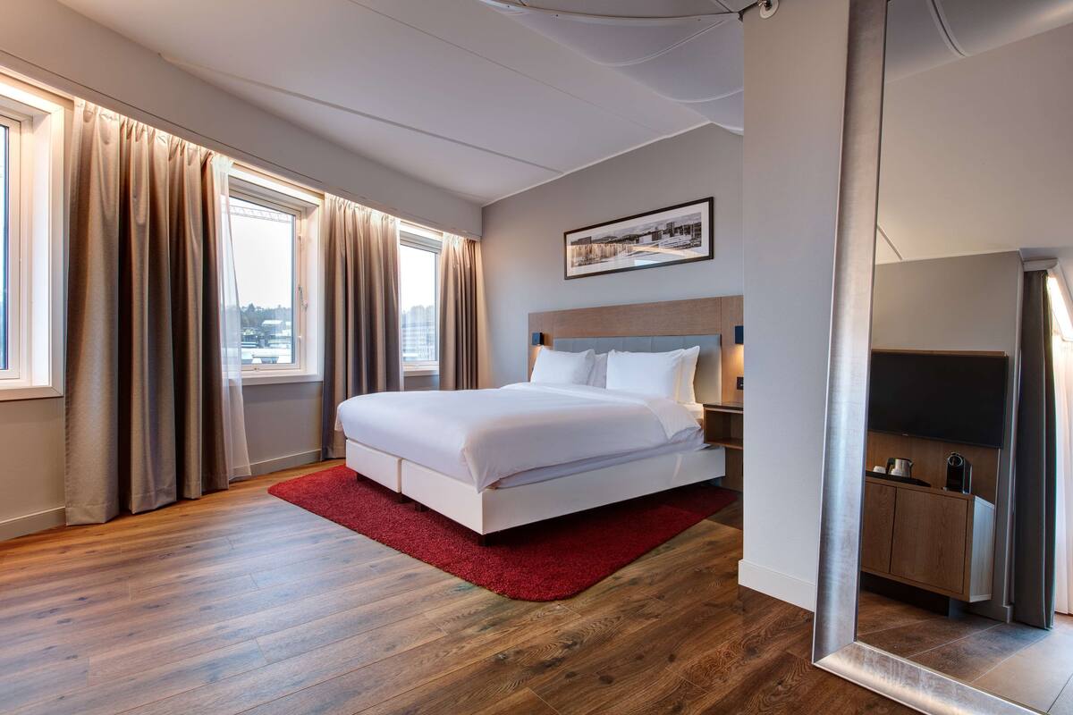 Quarto de hotel moderno com cama grande, piso de madeira, tapete vermelho, vista para a cidade e televisão na parede.
