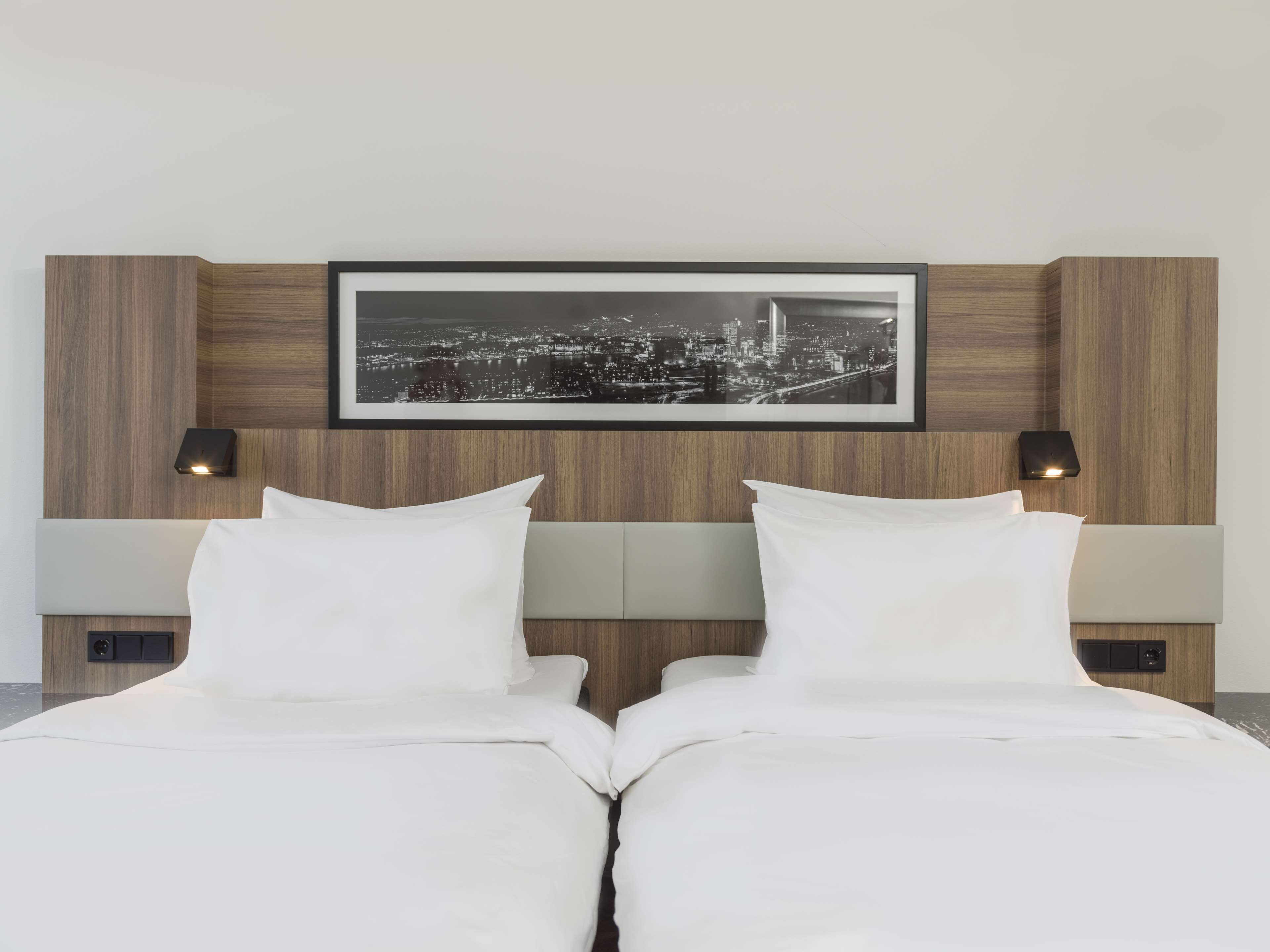 A hotel room with a large bed, two bedside tables, a framed cityscape picture, and wall-mounted reading lights.