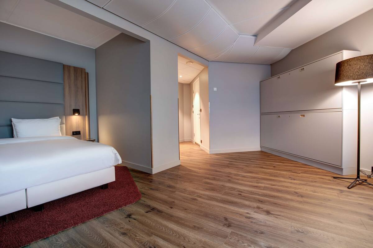 Quarto de hotel moderno com uma cama grande, um tapete vermelho, piso de madeira, uma mesa de cabeceira, uma luminária e um guarda-roupa.