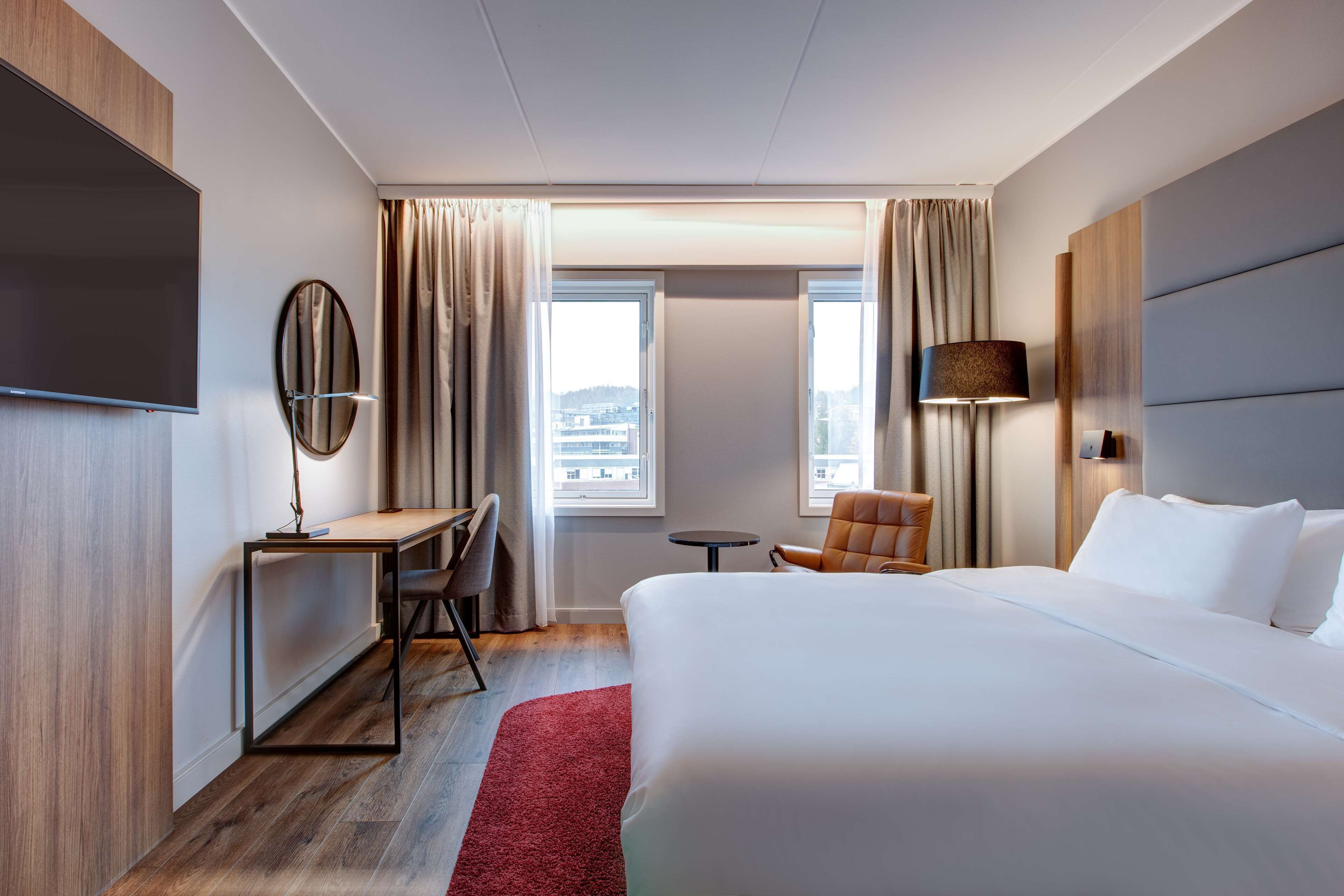 Habitación de hotel con una cama grande, un escritorio con silla, televisión, lámpara y vistas a la ciudad desde las ventanas.