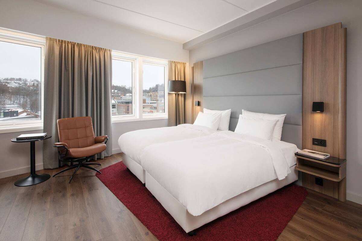 Quarto de hotel moderno com uma cama grande, uma poltrona, uma mesa pequena e vista para a cidade pela janela.