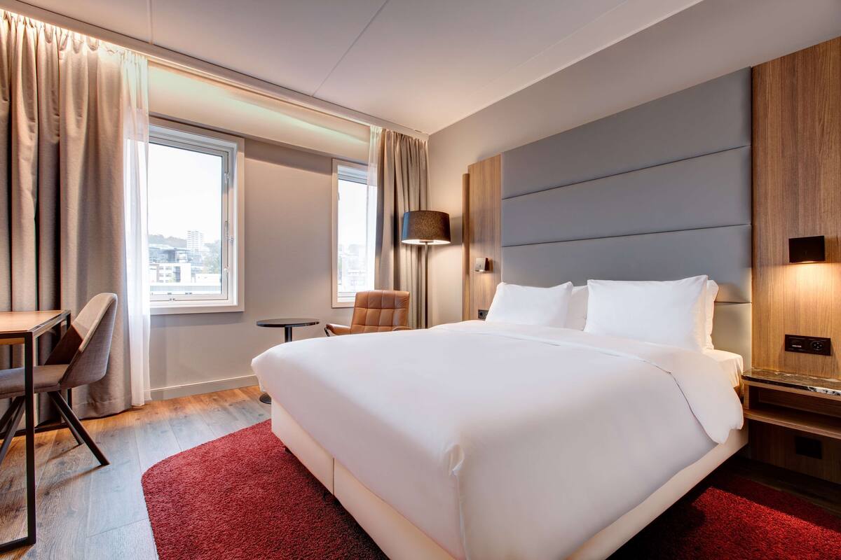 Quarto de hotel com uma cama grande, uma escrivaninha, uma cadeira, um abajur e vista para a cidade pela janela.