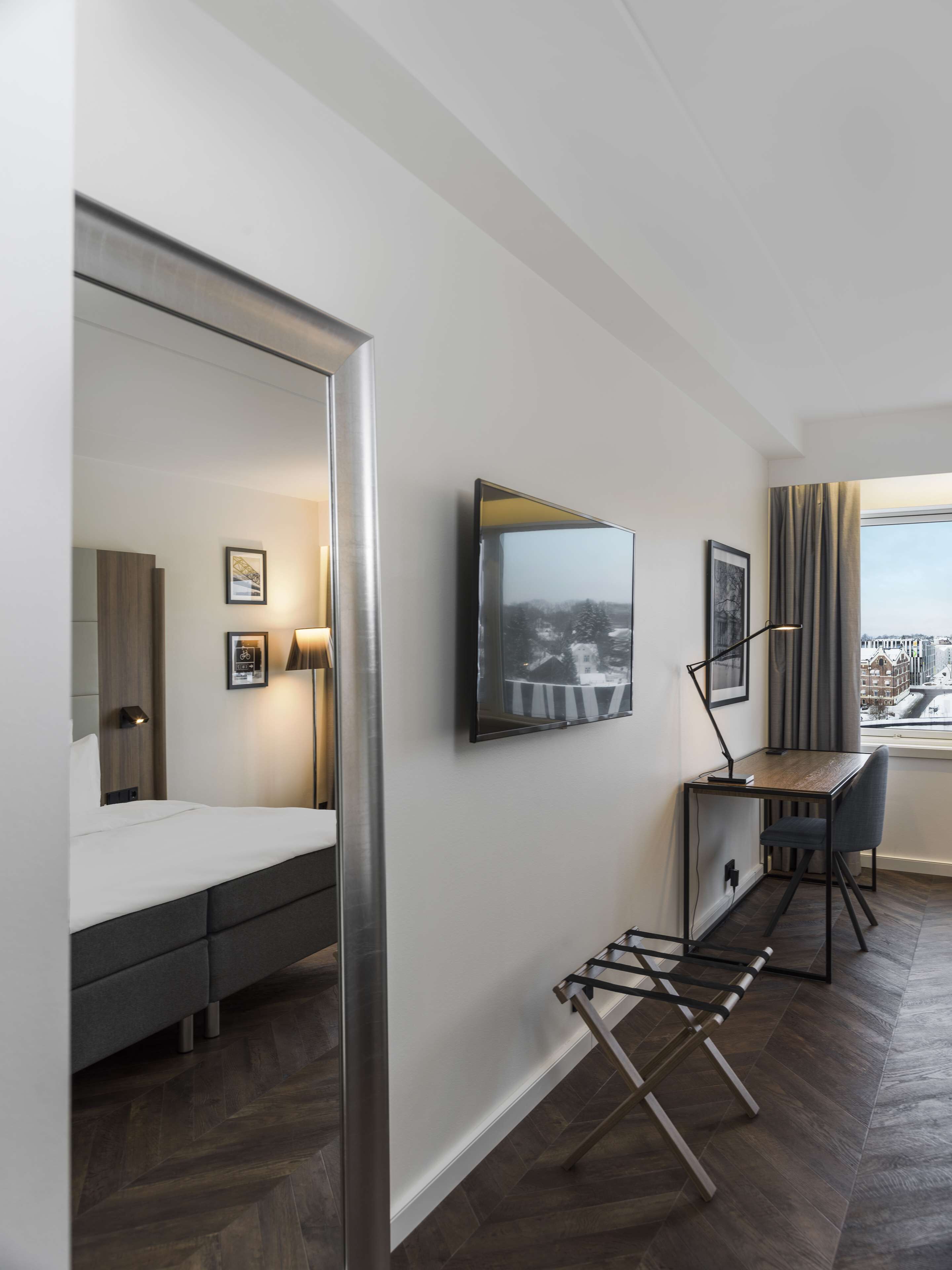 A modern hotel room with a bed, a desk, and a chair. There are two framed pictures on the wall, a lamp, and a window with a view of buildings.