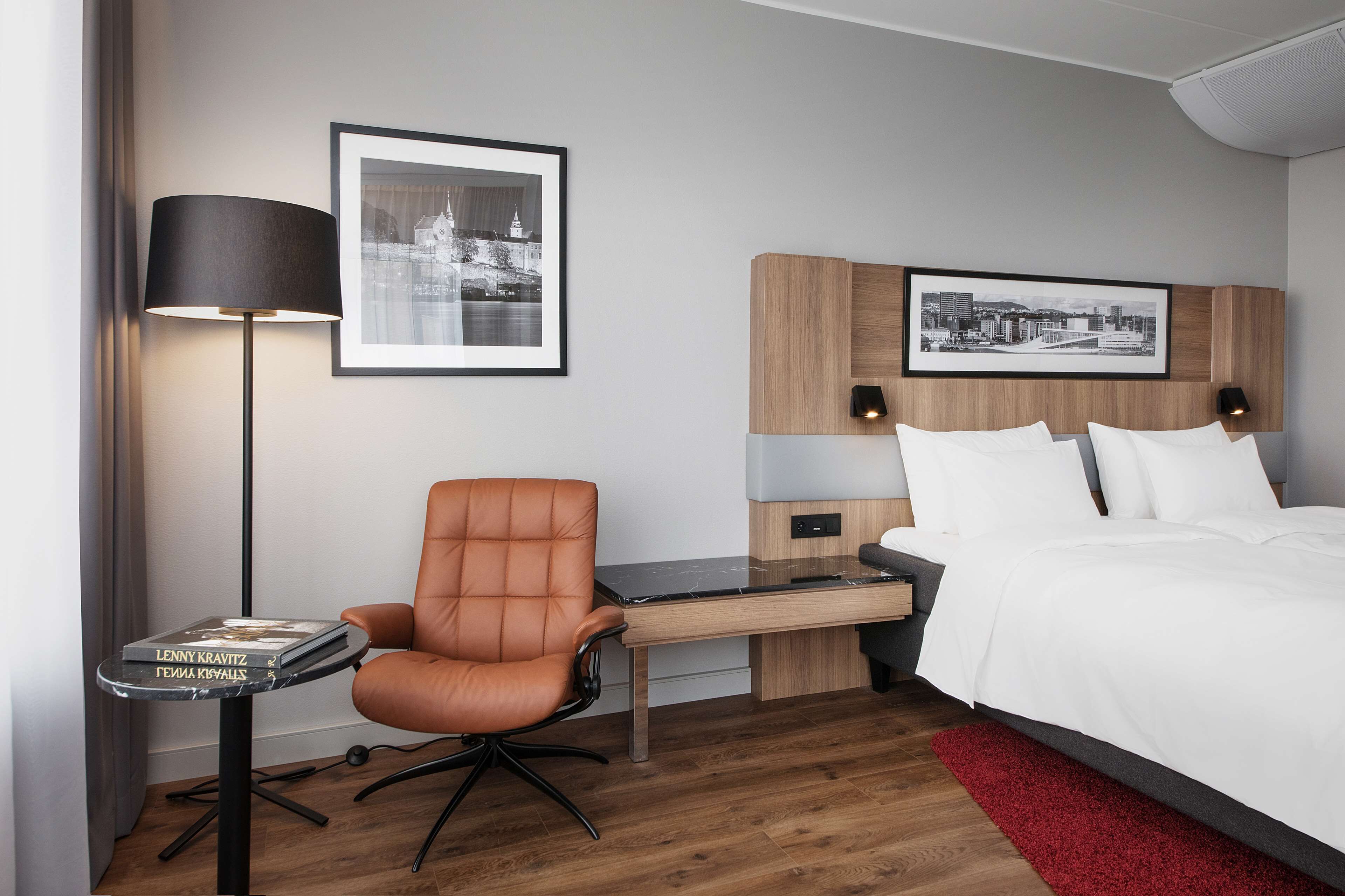 A hotel room with a bed, a desk with a chair, a lamp, and two framed pictures on the wall.