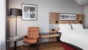 A hotel room with a bed, a desk with a chair, a lamp, and two framed pictures on the wall.