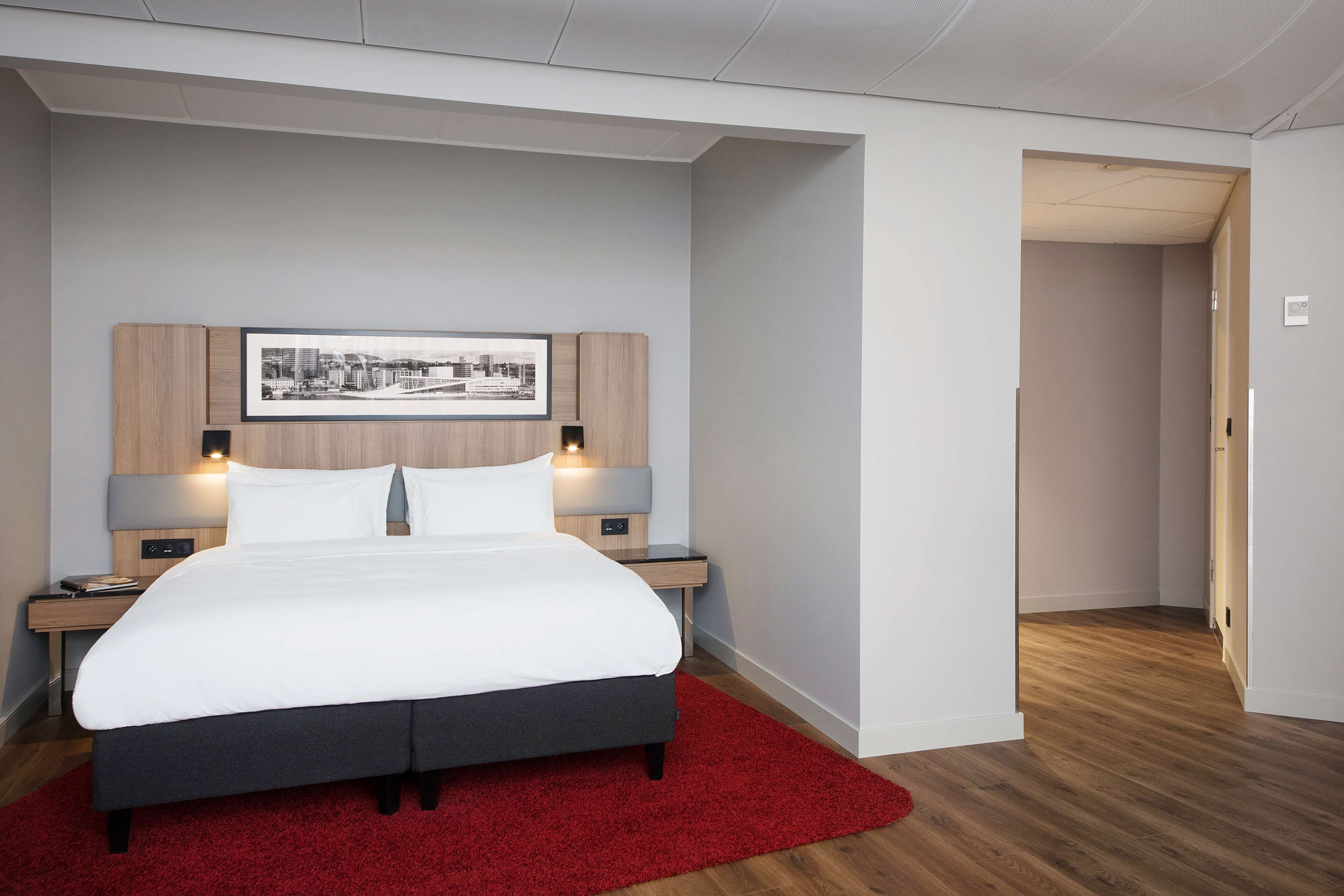 Une chambre à coucher moderne avec un grand lit, une tête de lit en bois, deux tables de chevet avec des lampes, un tapis rouge et un tableau représentant un paysage urbain encadré accroché au mur.