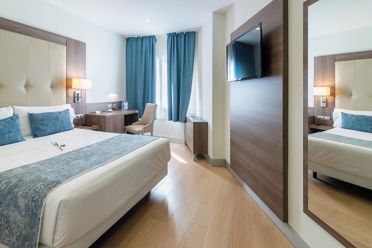 A hotel room with a large bed, a desk, a chair, a TV, and a mirror.