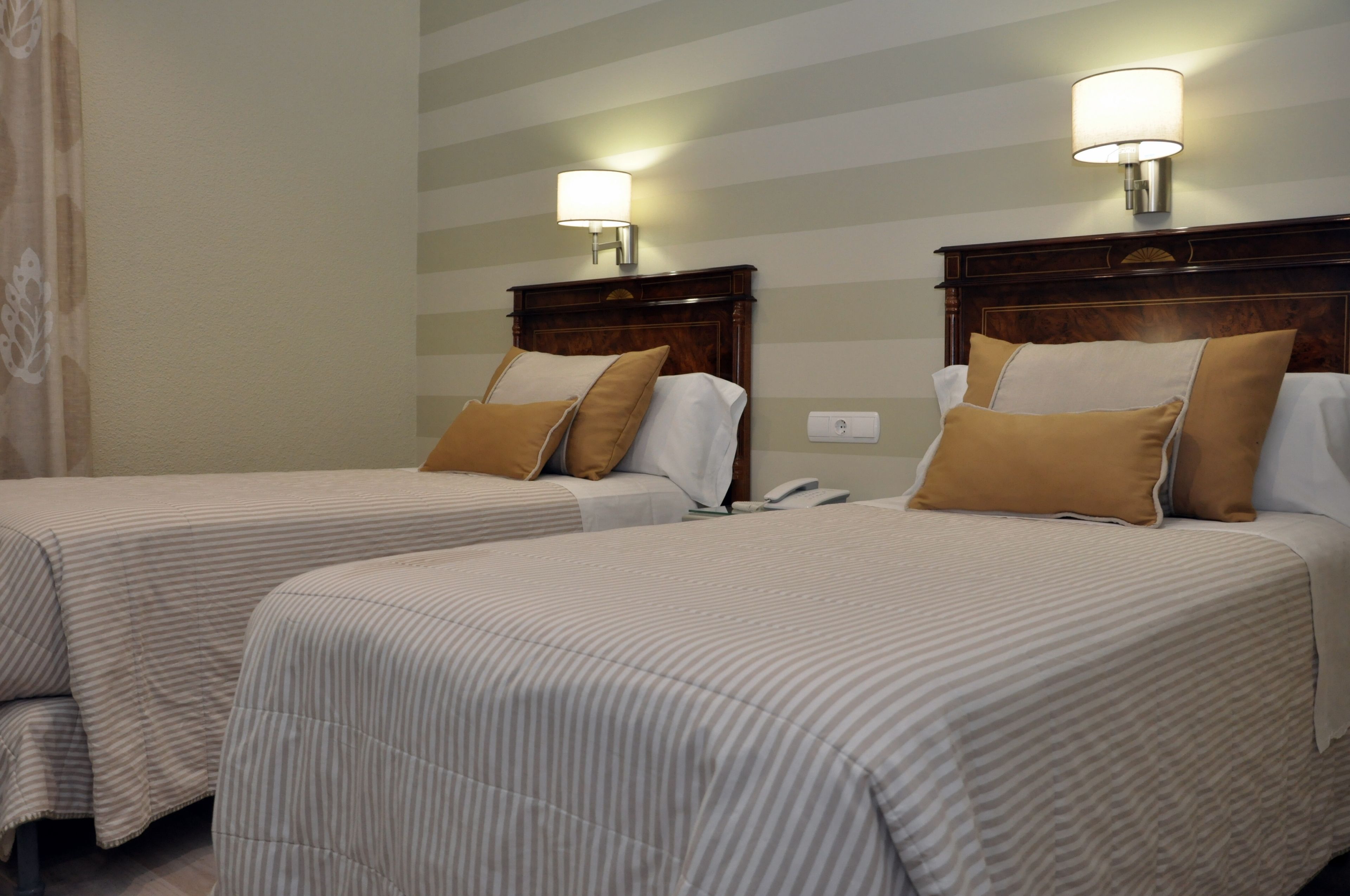 A hotel room with two beds, striped bedding, a wooden headboard, a nightstand, and a wall-mounted lamp.