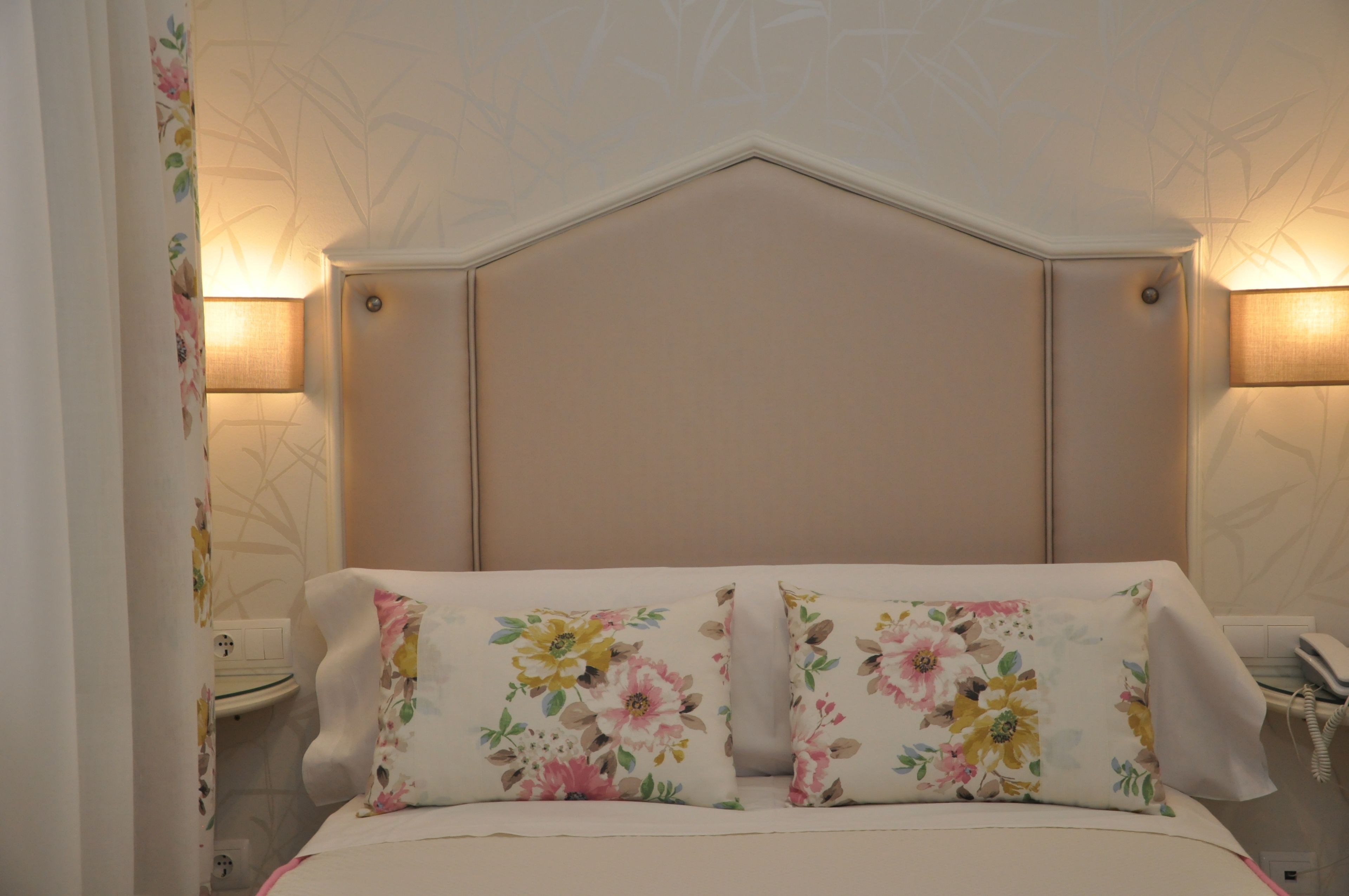 Un dormitorio con cabecera, dos lámparas de noche y almohadas estampadas con motivos florales.