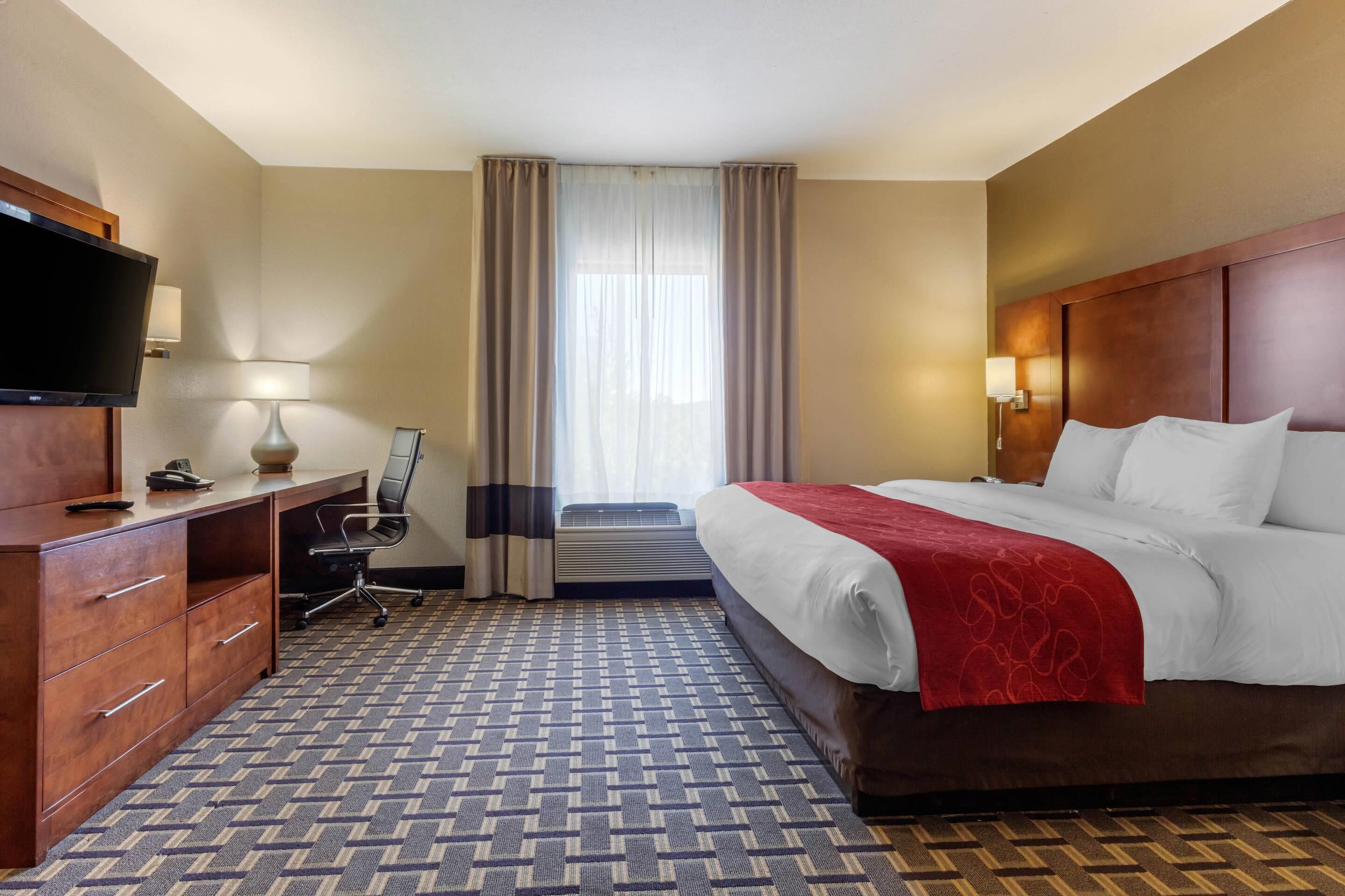 Une chambre d’hôtel avec un lit, un bureau avec un ordinateur, une télévision et une fenêtre avec des rideaux.