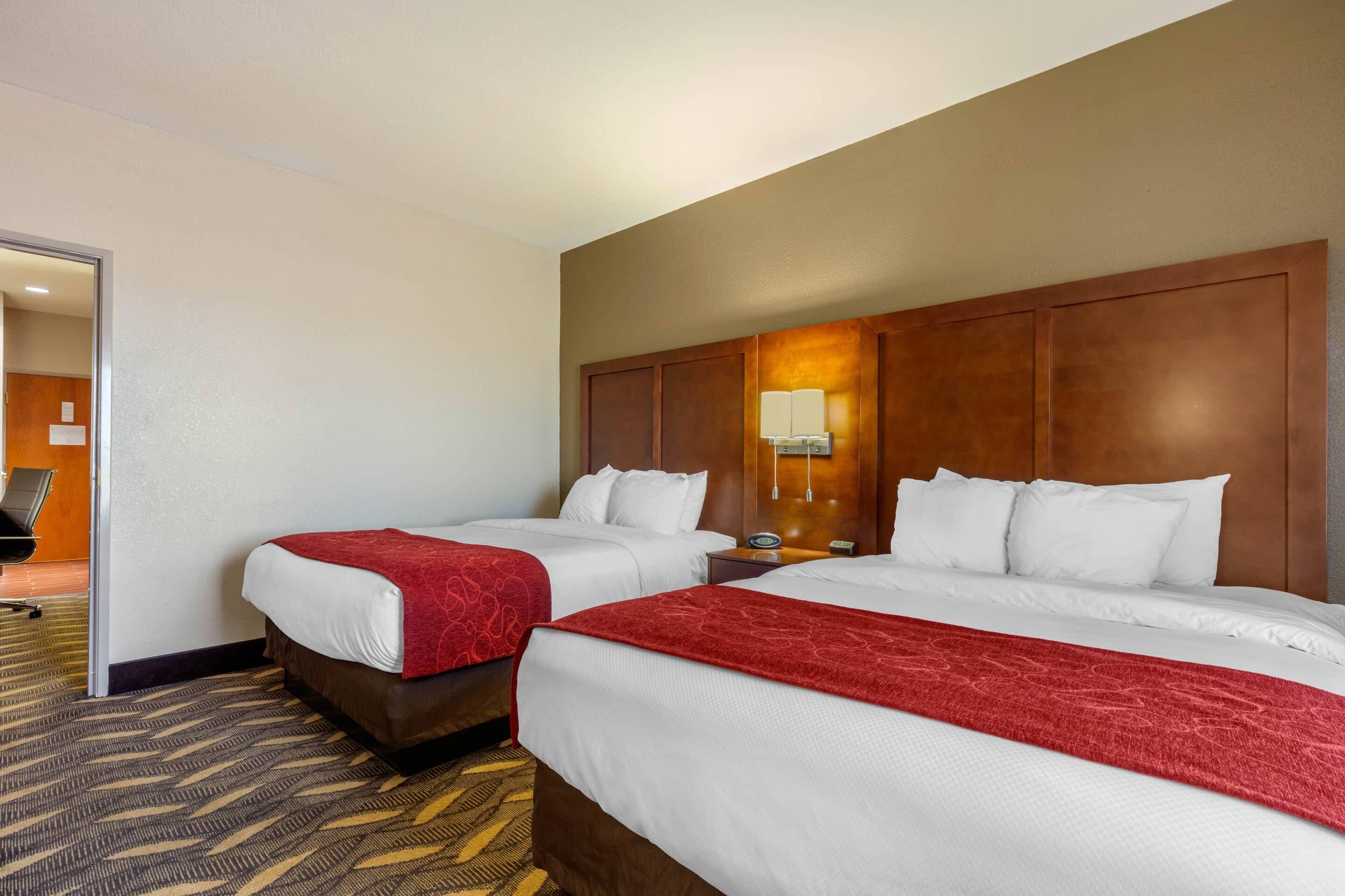 Une chambre d’hôtel avec deux lits, une tête de lit en bois, une table de chevet avec une lampe et un sol recouvert de moquette.