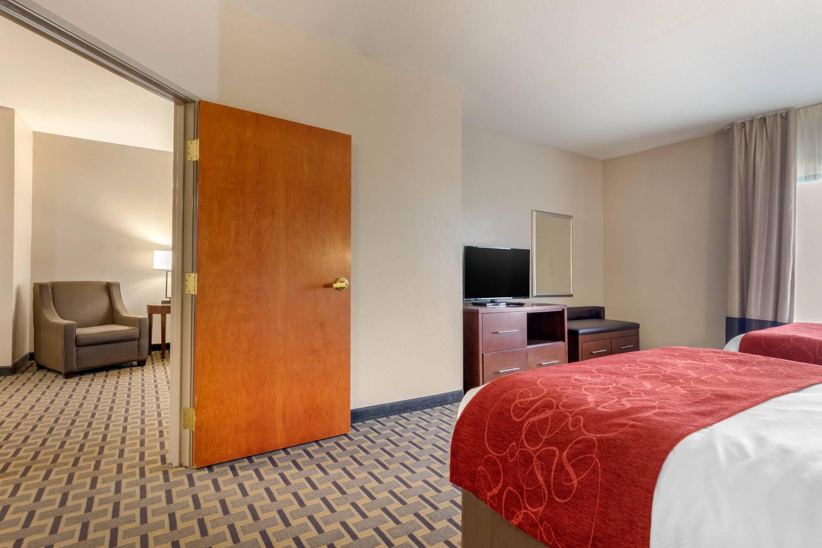 A hotel room with a patterned carpet, a wooden door, a television on a dresser, a chair, and two beds with red bedspreads.