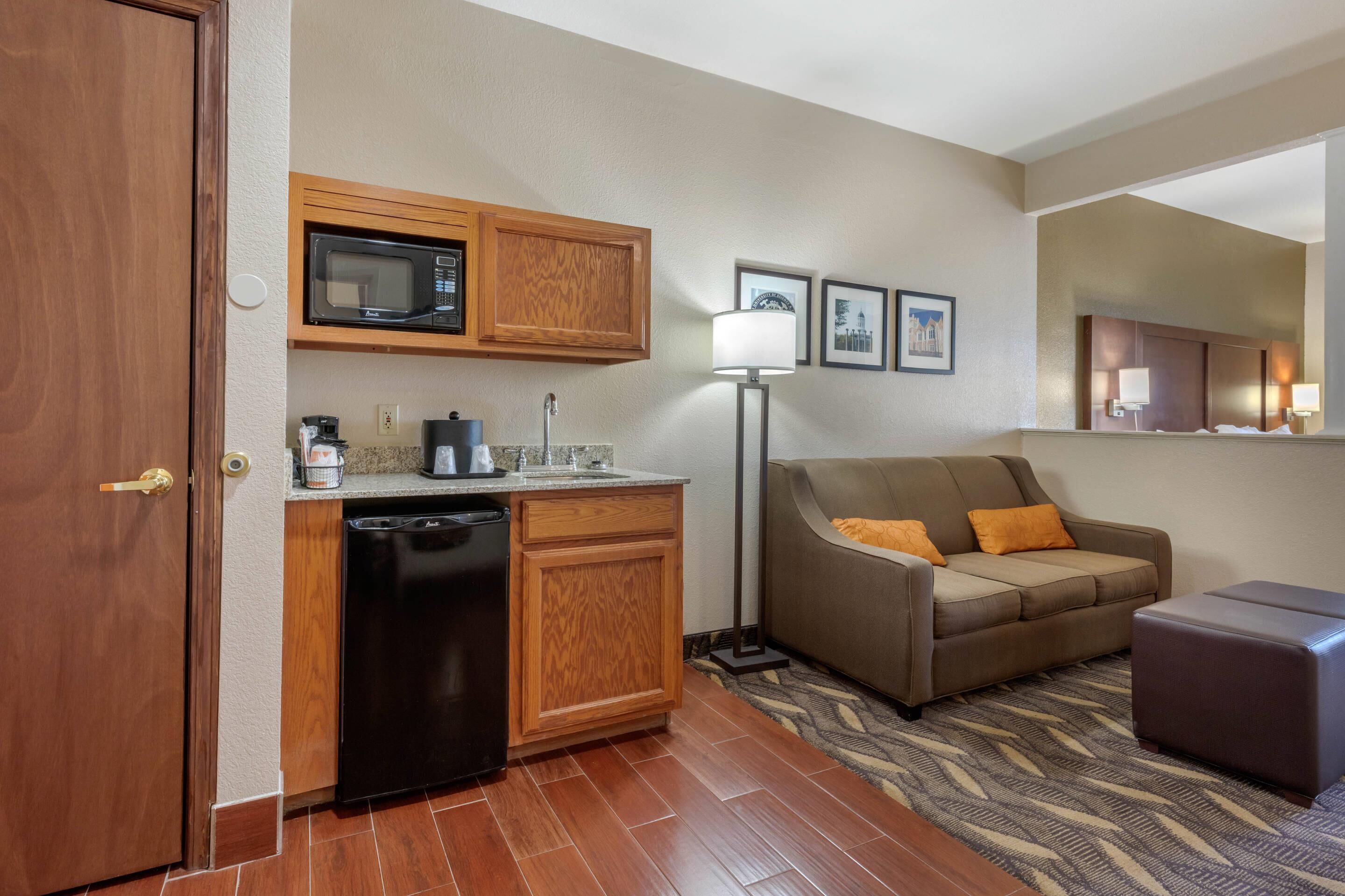 A compact hotel room with a kitchenette, a sofa, a small ottoman, and a floor lamp.