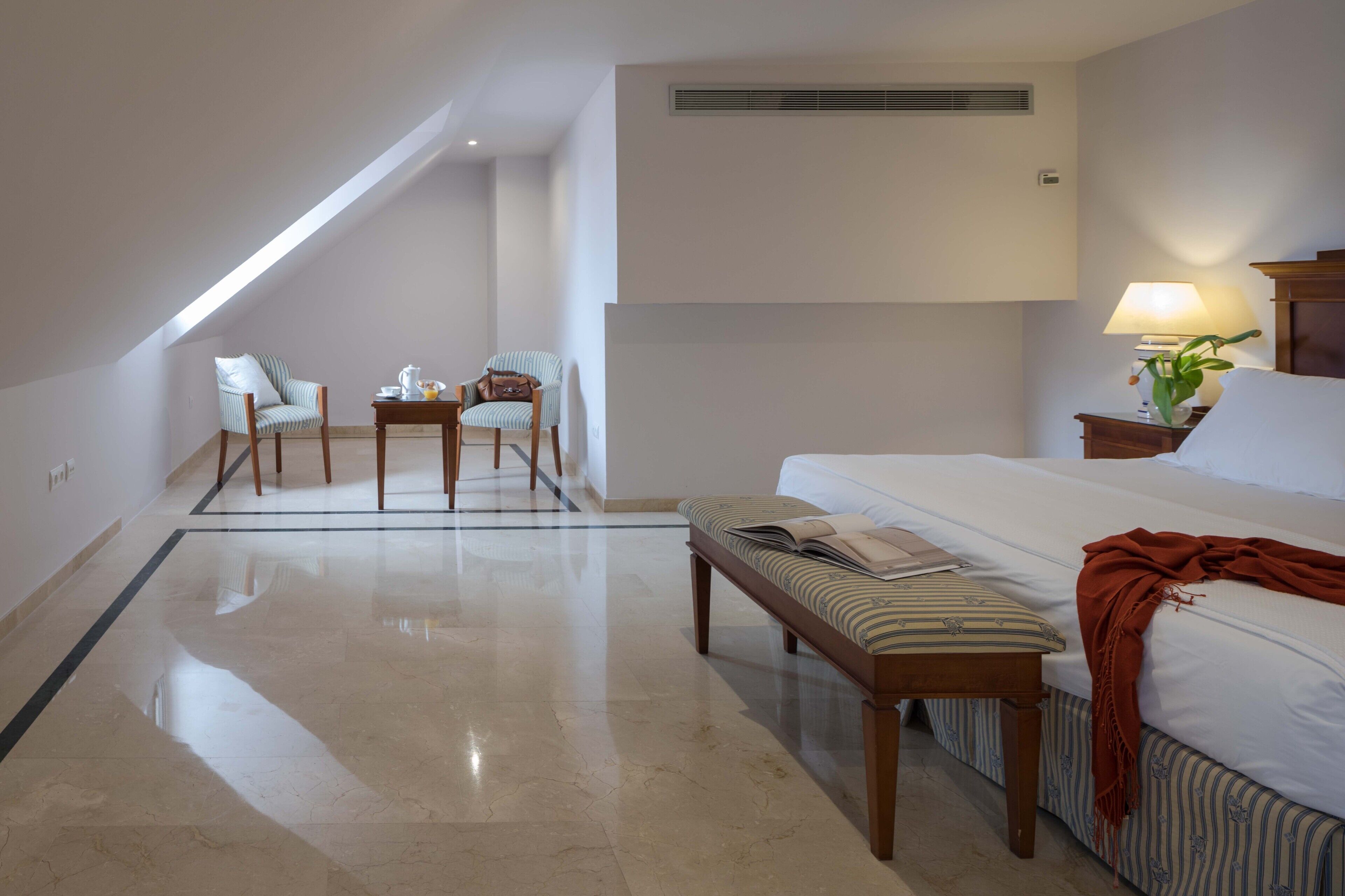 A hotel room with a bed, a bench, a small table with chairs, and a lamp.