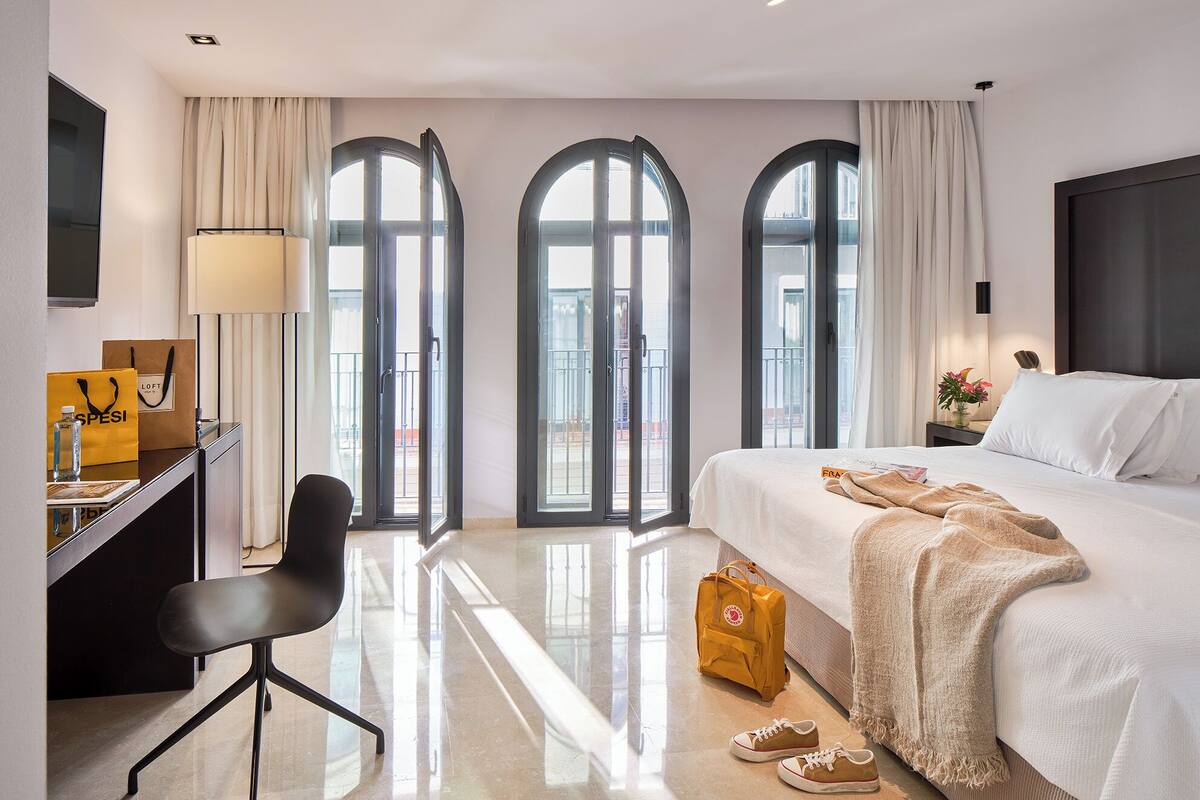 A hotel room with a bed, a desk with a chair, a lamp, and a view through large arched windows.