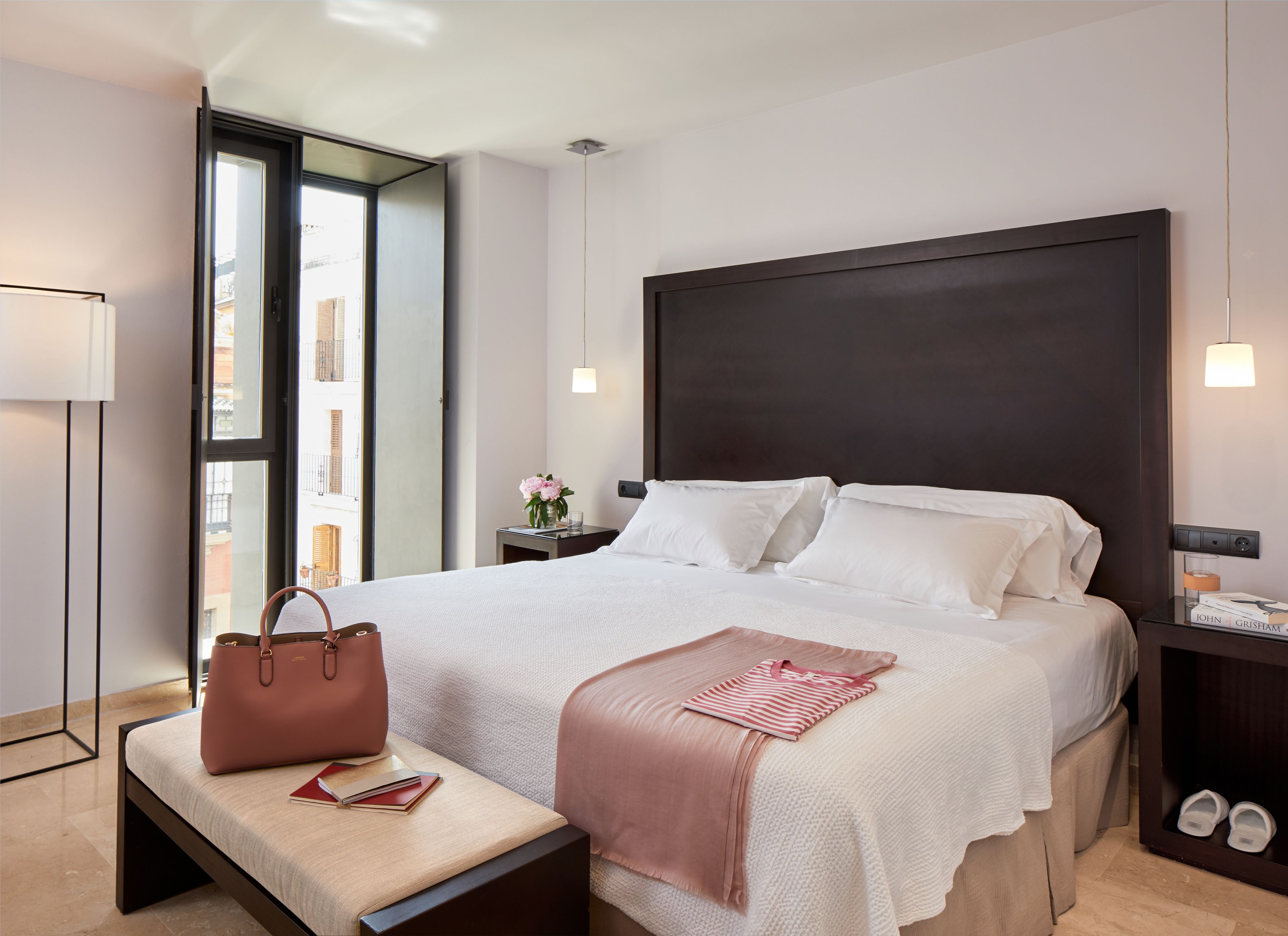 A modern hotel room with a large bed, bedside tables, a bench, and a view of the city.