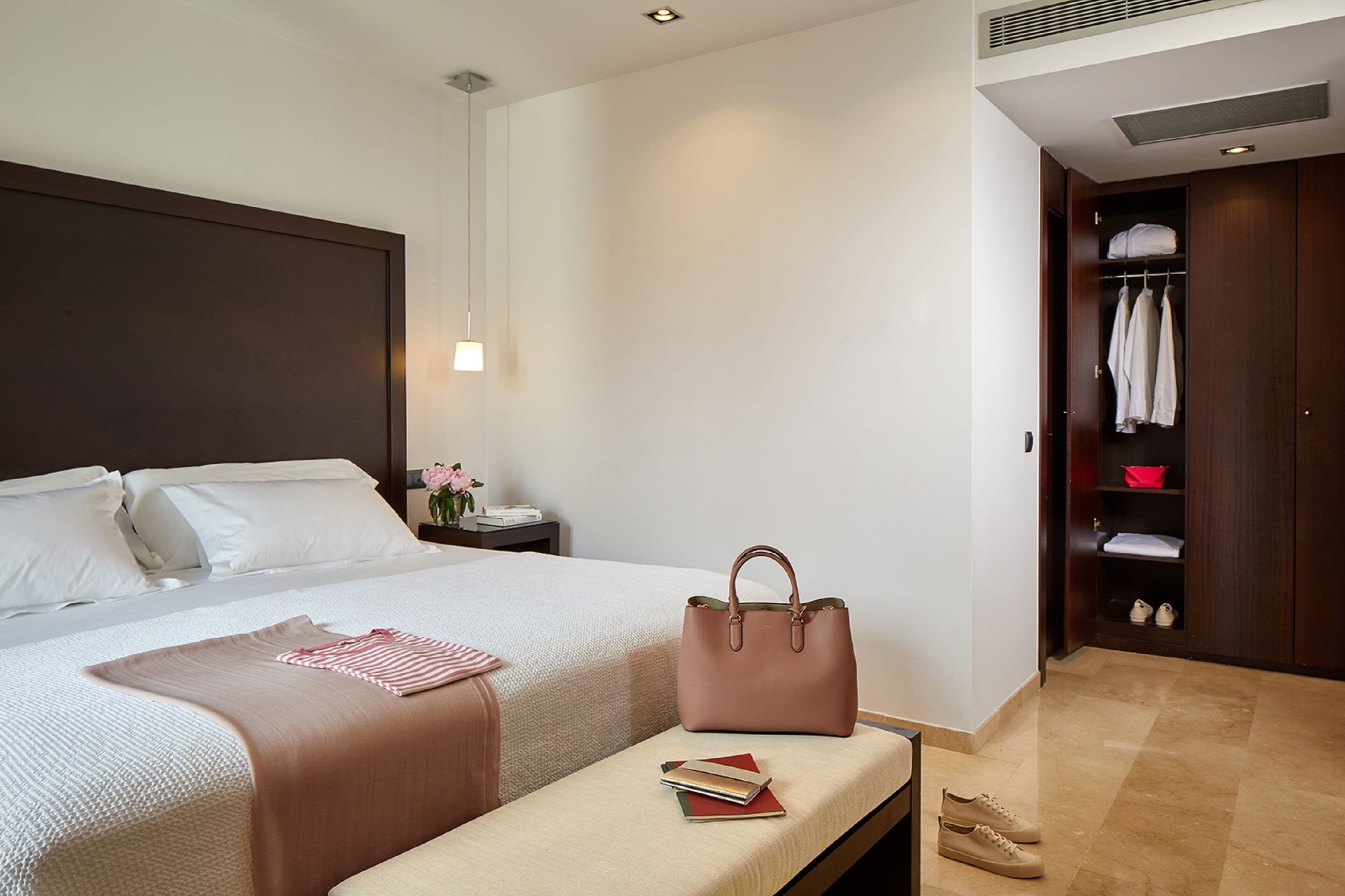 A hotel room with a bed, bedside table, lamp, bench, and a wardrobe with hanging clothes and shoes.