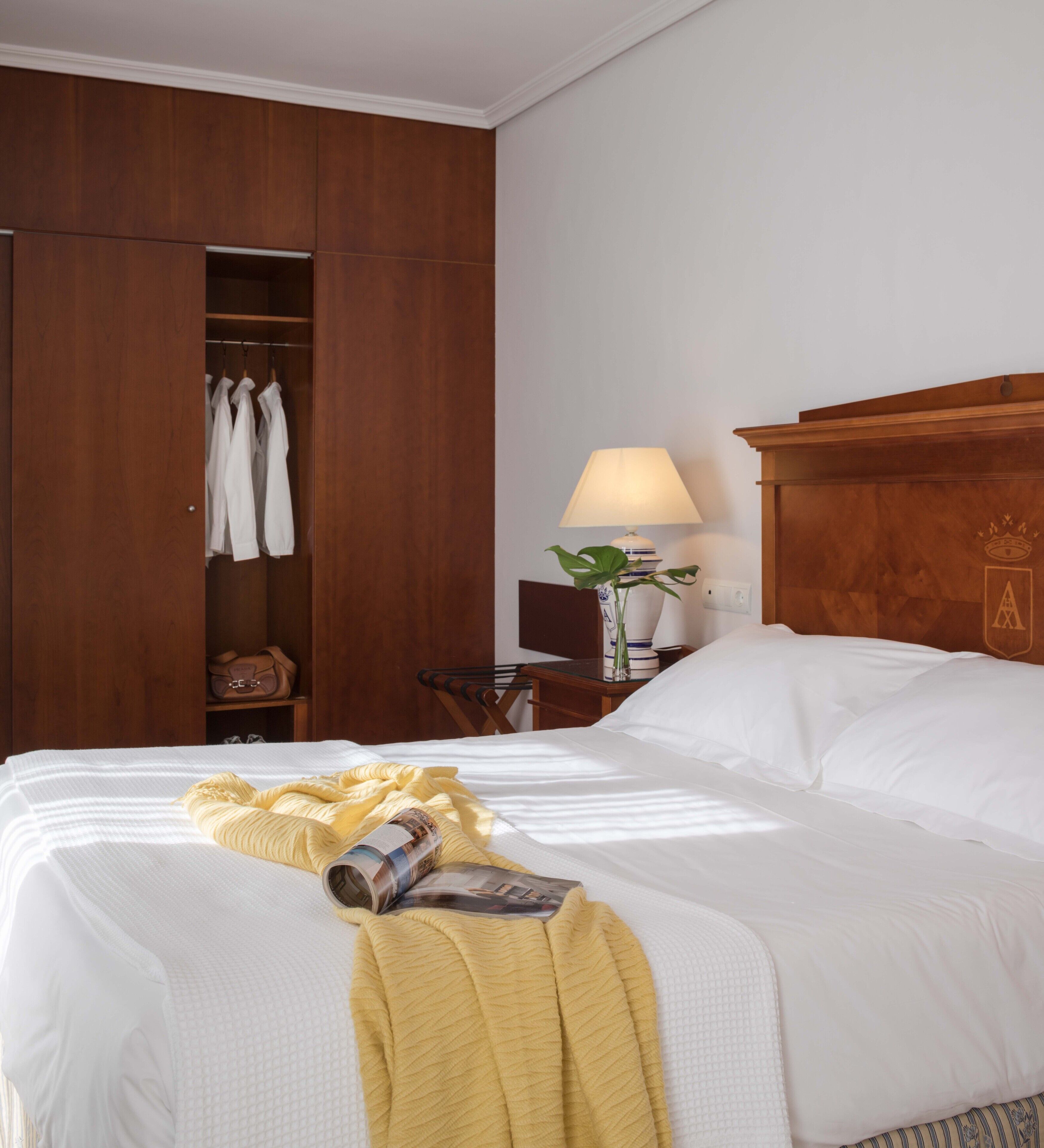A neatly made bed with a yellow blanket and a magazine, a wooden wardrobe with hanging clothes, a bedside table with a lamp and a vase, and a wooden headboard with a crest.