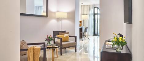 Une chambre d’hôtel moderne, dotée d’un canapé, d’un fauteuil et d’un bureau avec une lampe. Un grand miroir orne le mur, et un téléviseur à écran plat y est fixé.