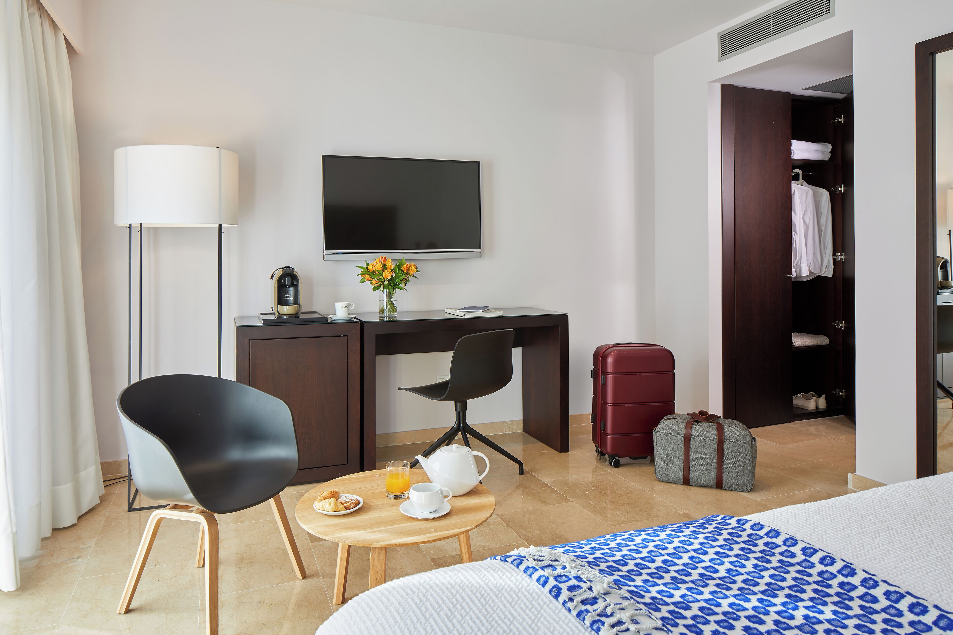 Habitación de hotel con cama, escritorio con silla, televisor, lámpara, mesita con artículos de desayuno y armario.