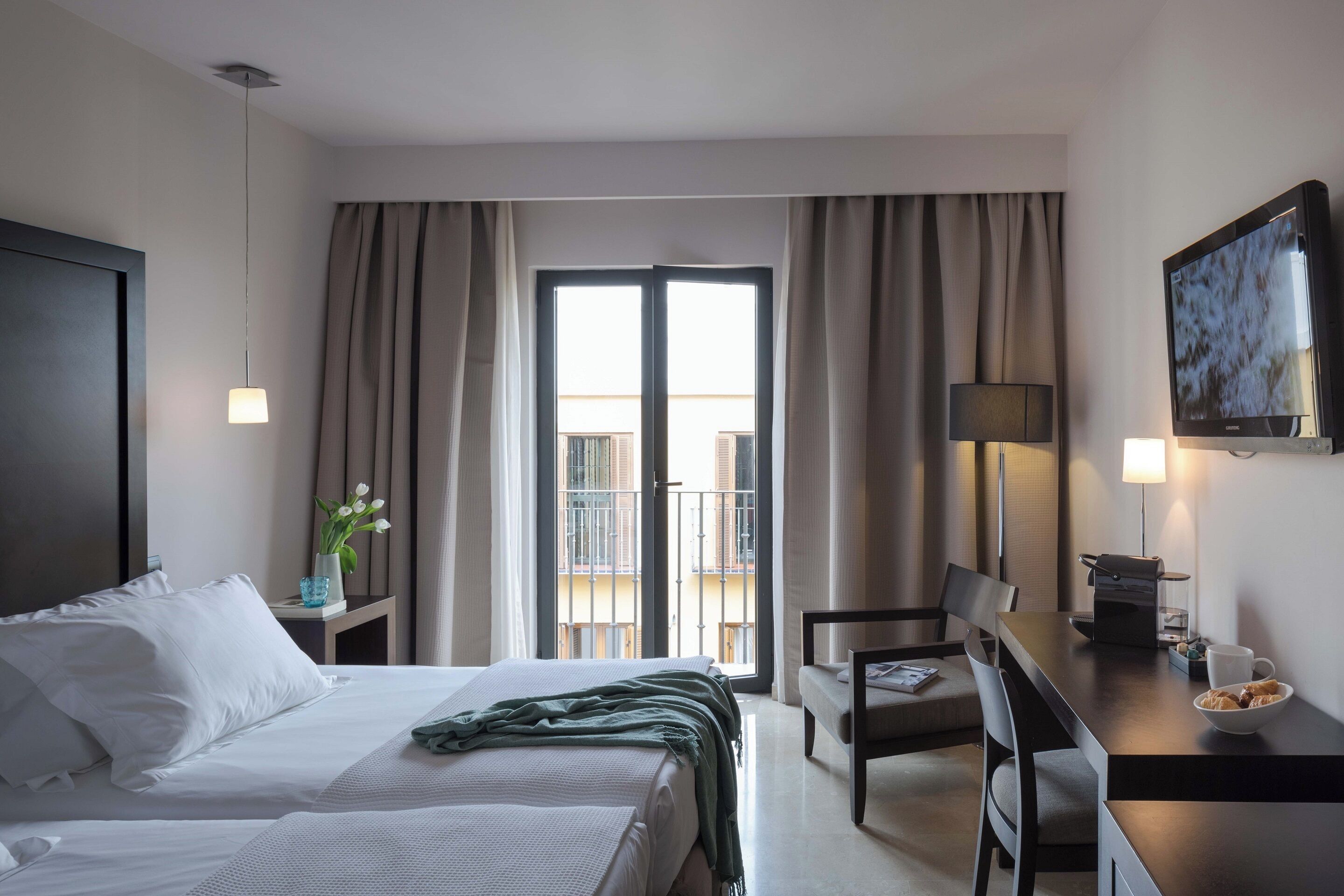 Habitación de hotel con cama, escritorio con lámpara, silla, cafetera y televisor en la pared.