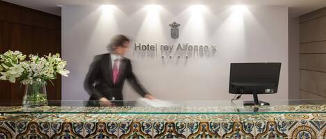 A hotel reception desk with a tiled pattern, a monitor, and a vase of flowers.