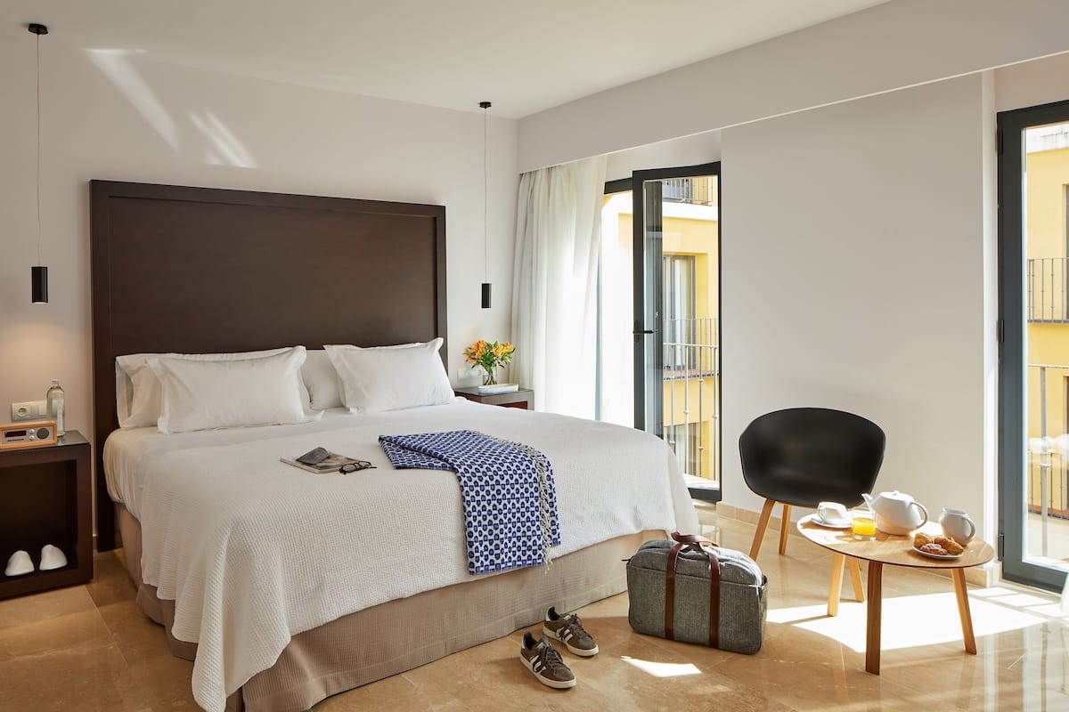 A hotel room with a large bed, a bedside table with a radio, a suitcase, a chair, and a breakfast table with a coffee pot and orange juice.