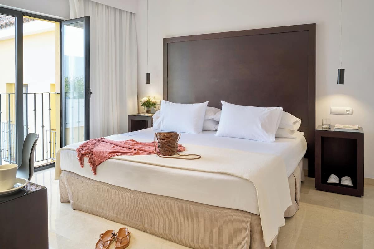 A hotel room with a large bed, a bedside table with a lamp, a small table with a cup and saucer, and a pair of shoes on the floor.
