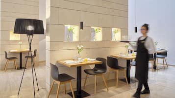 Un área de comedor moderna con mesas de madera y sillas negras, una lámpara de pie negra y un camarero con uniforme.