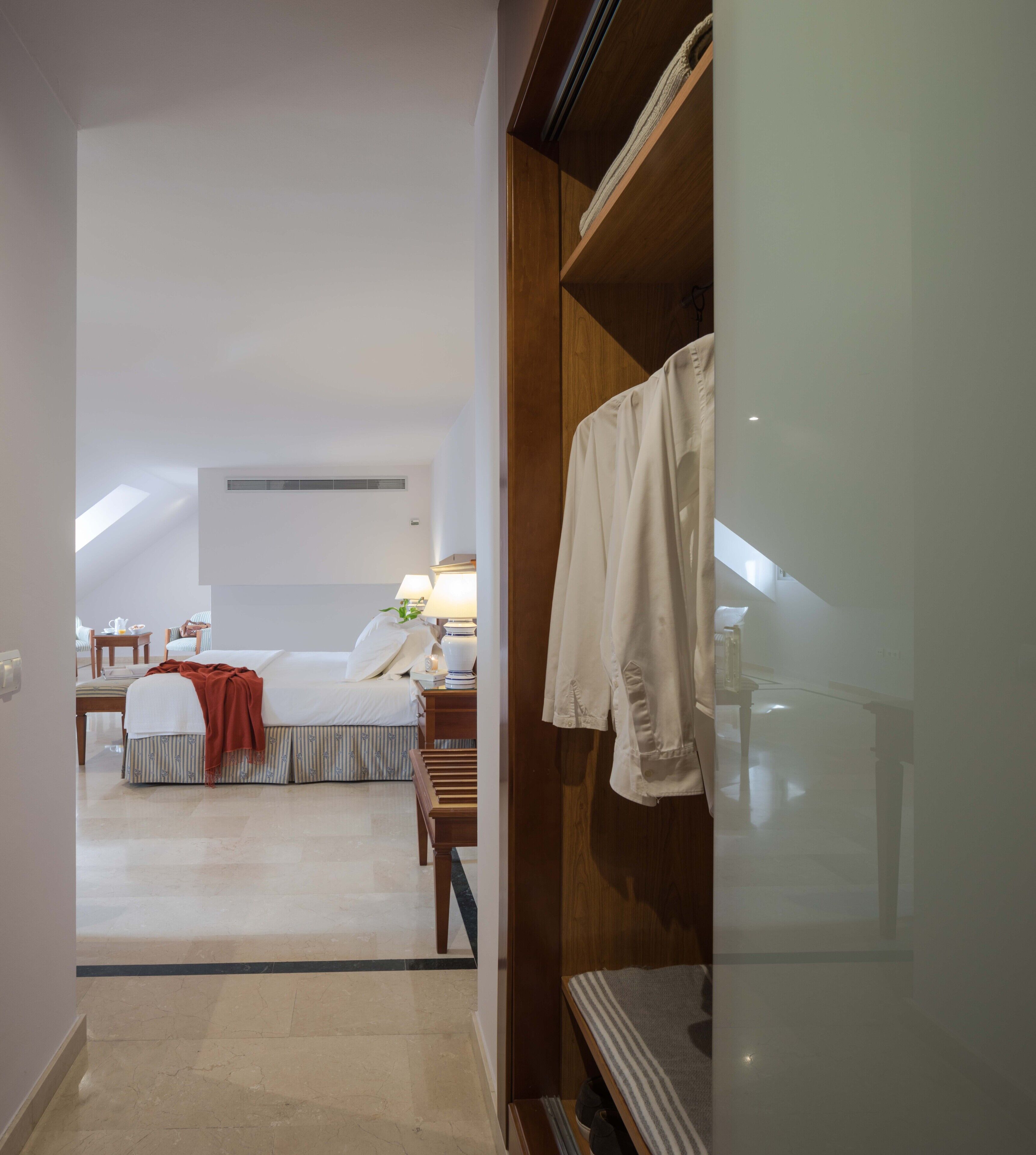 A hotel room with a bed, bedside table, lamp, and a view of the adjacent room.