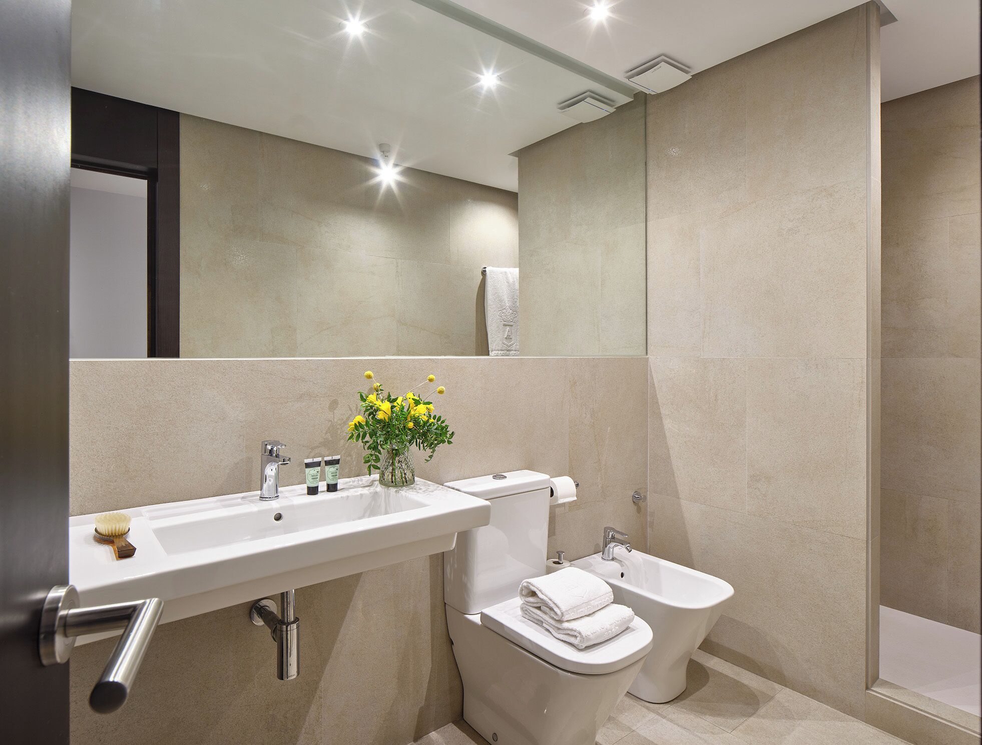 A modern bathroom with a white sink, toilet, and bidet, a mirror, and a vase with yellow flowers.