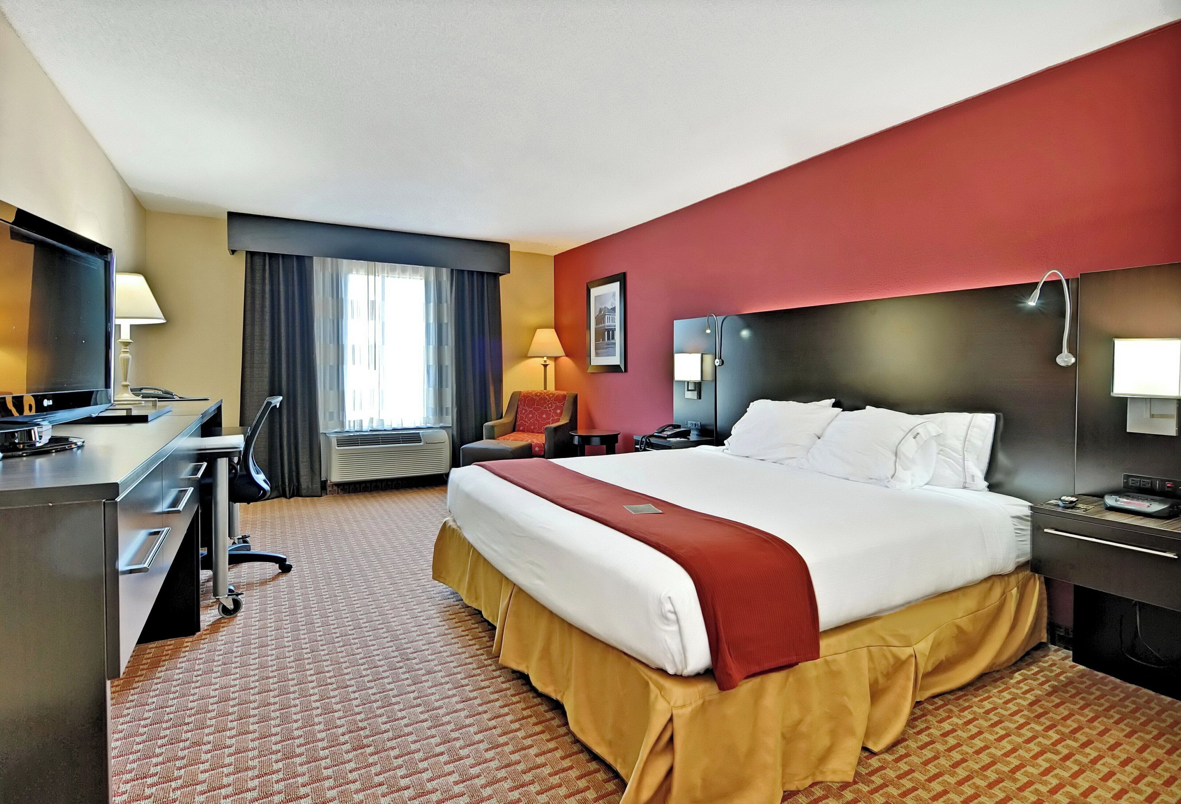 A hotel room with a large bed, a desk, a chair, a TV, and a window with curtains.