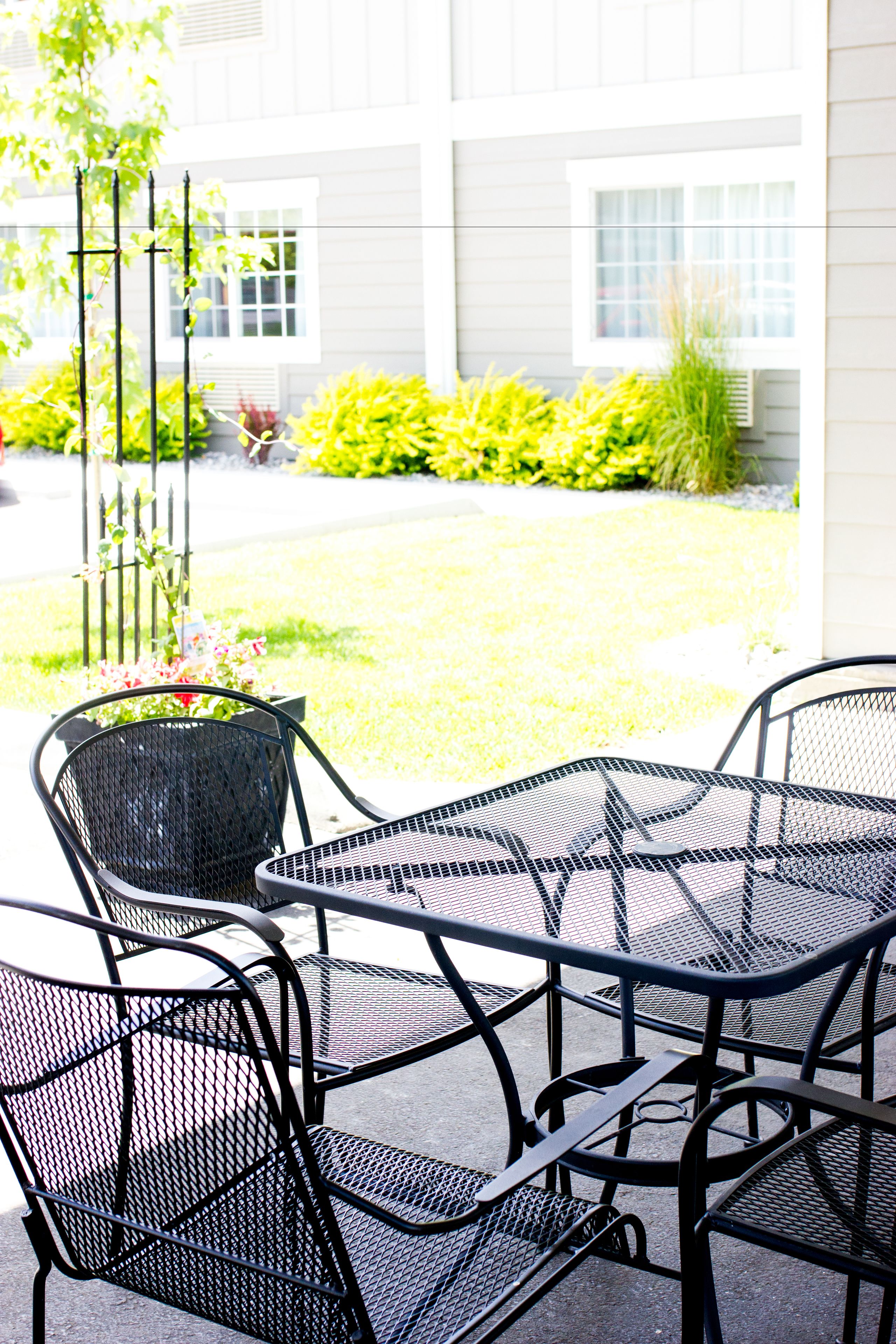 A patio area with a metal table and chairs, surrounded by a garden with various plants and a building in the background.