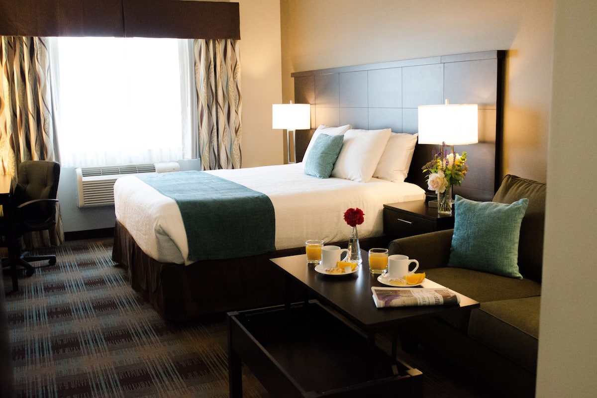 Habitación de hotel con una cama grande, dos lámparas de noche, una mesa de centro con alimentos para el desayuno y vistas de la ciudad a través de la ventana.