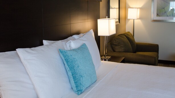 A hotel room with a large bed, a nightstand with a lamp, a chair, and a framed picture on the wall. - Inn At Moses Lake with Extended Stay (Moses Lake)