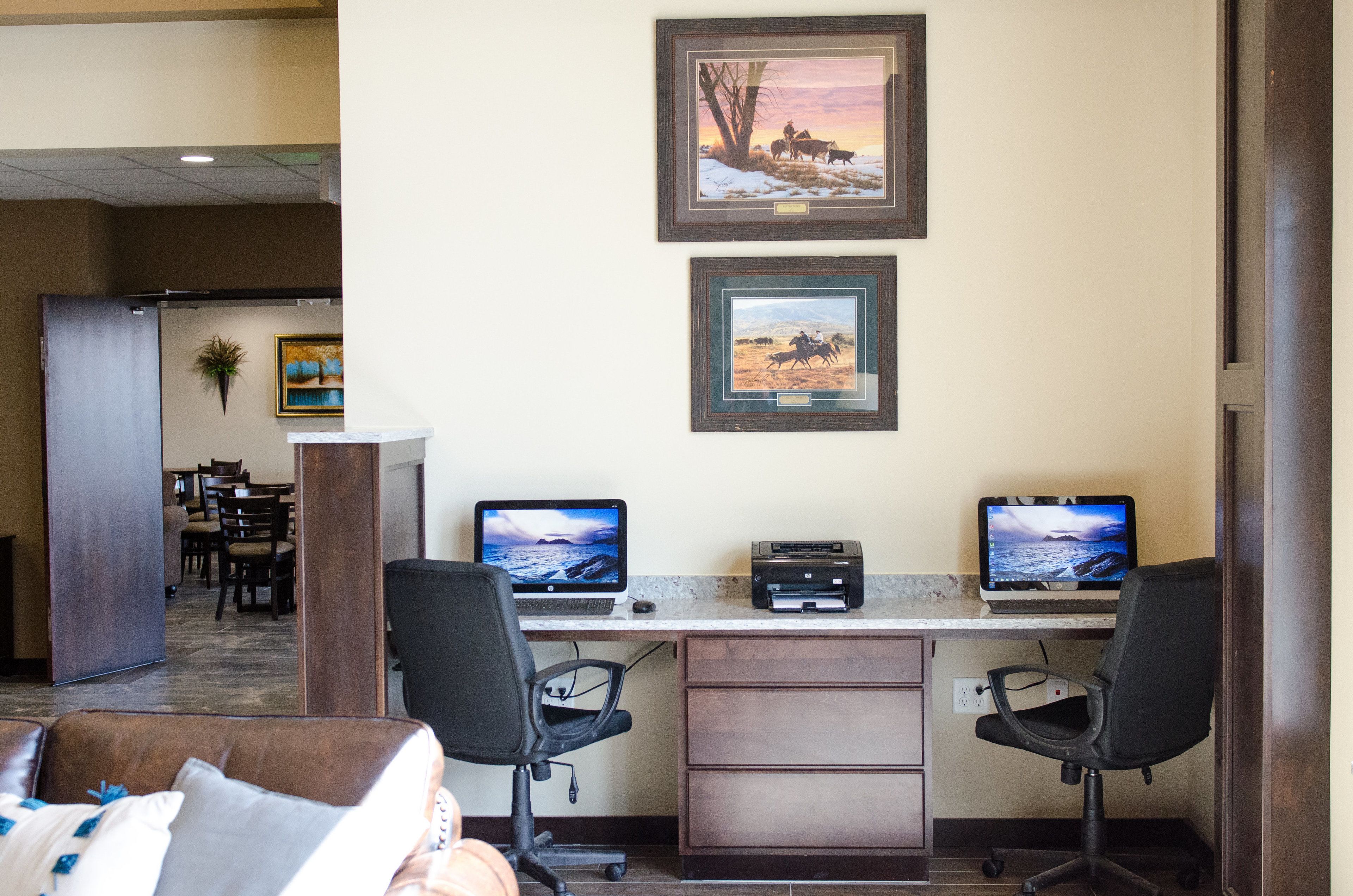 A hotel room with a desk, two chairs, a laptop, and a desktop computer. There is a framed picture on the wall and a plant on the desk.