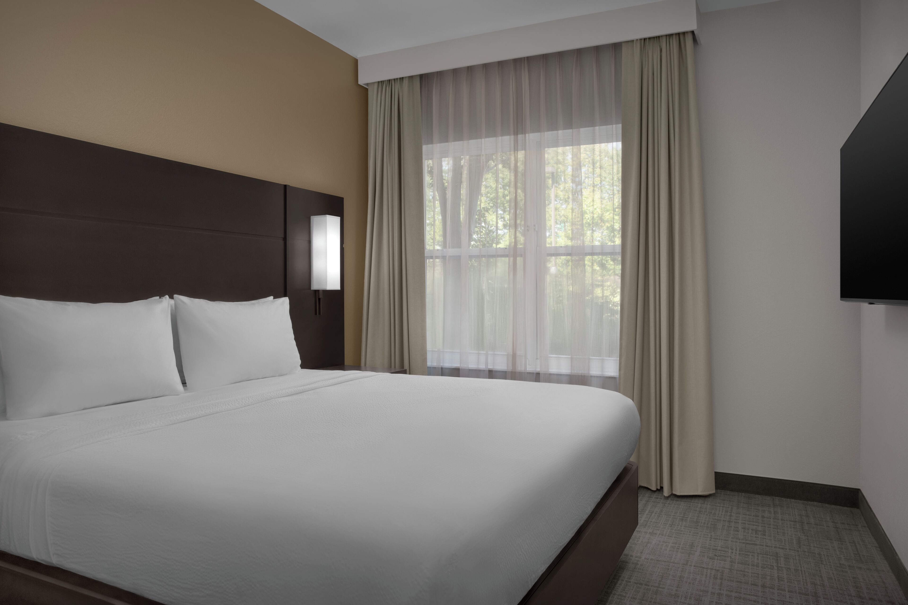 A hotel room with a large bed, a headboard, a TV, and a window with curtains.