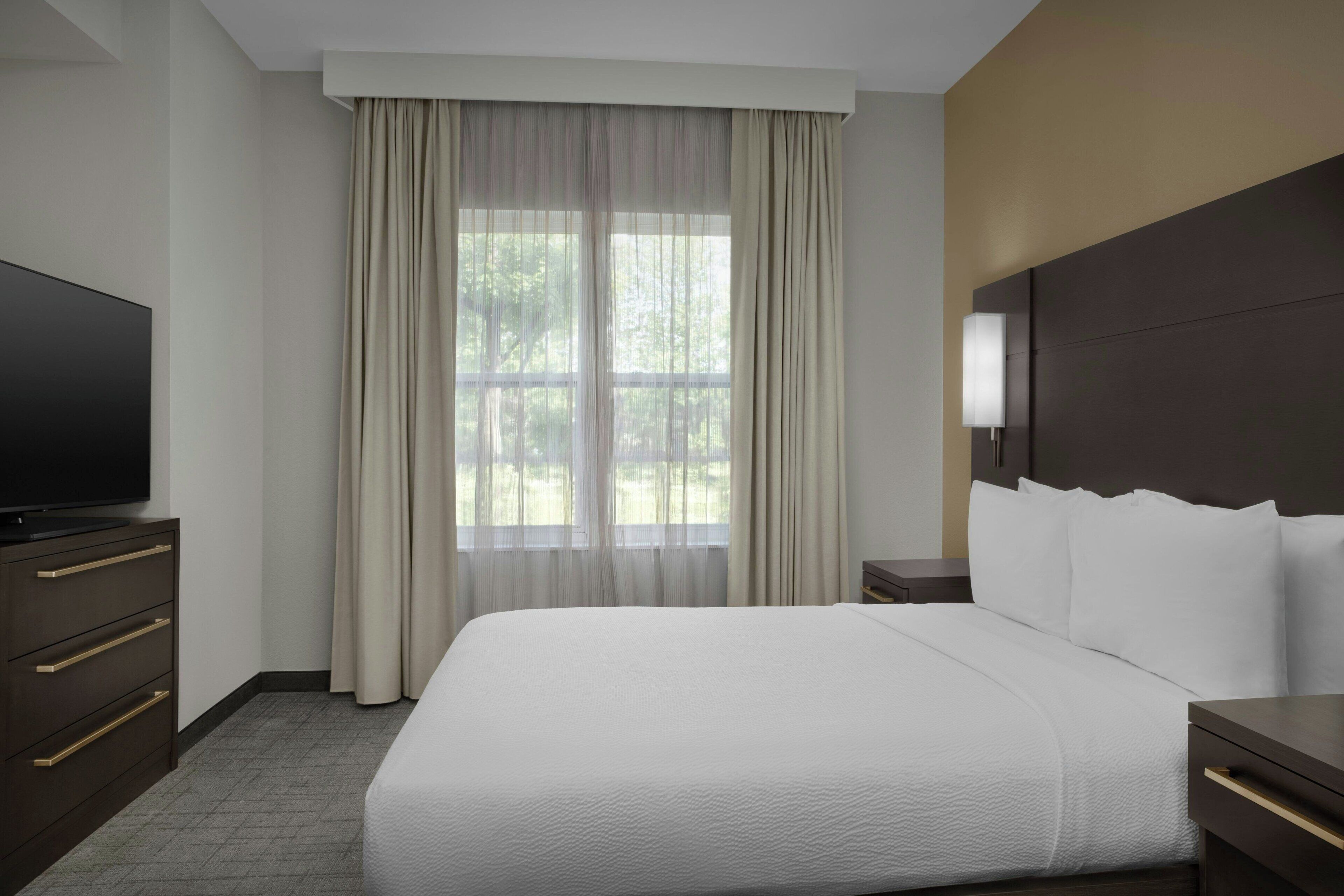 A hotel room with a large bed, a television on a dresser, a window with curtains, and a nightstand with a lamp.