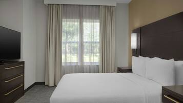 A hotel room with a large bed, a television on a dresser, a window with curtains, and a nightstand with a lamp.