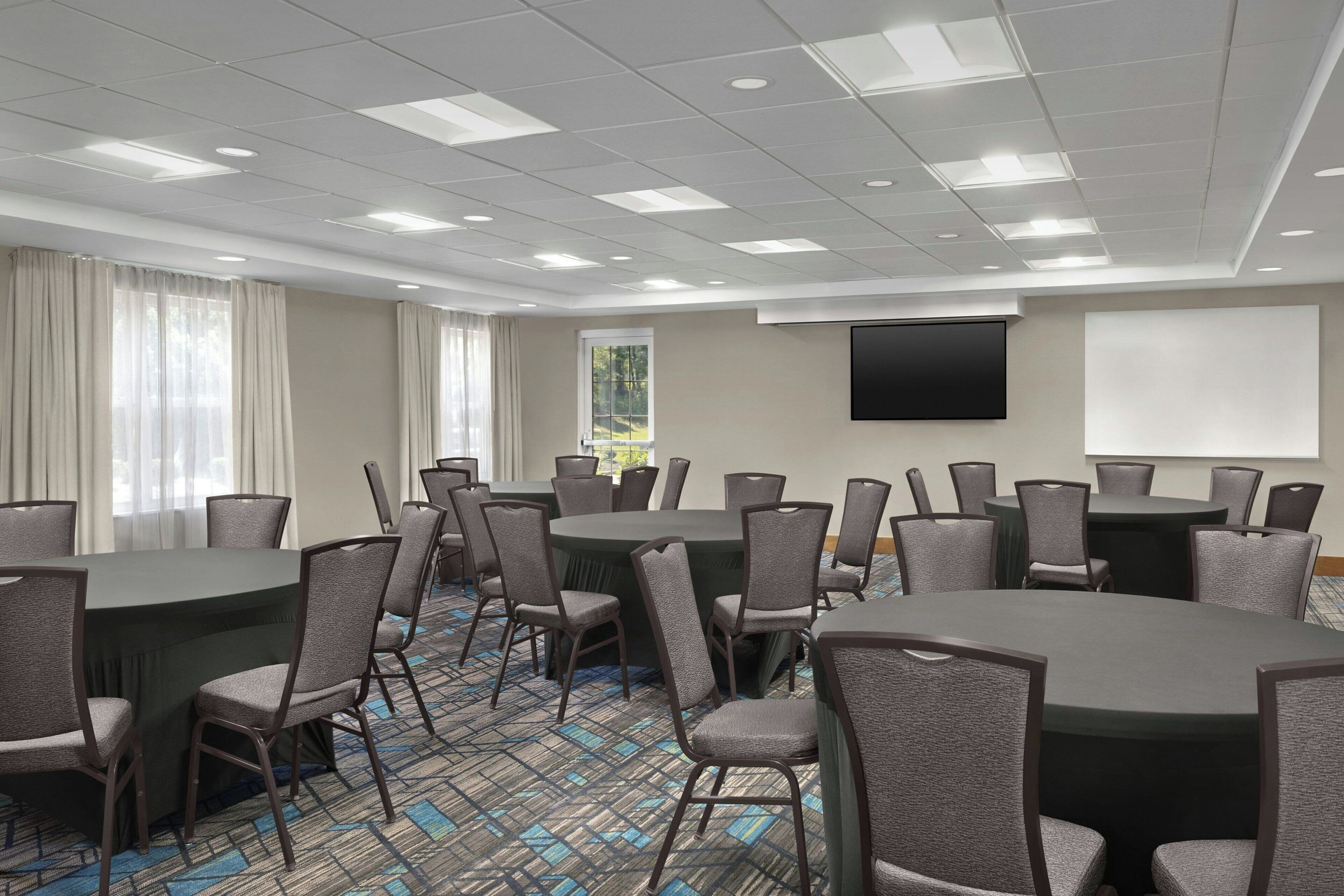 A conference room with round tables, chairs, a projector screen, and a large window with curtains.