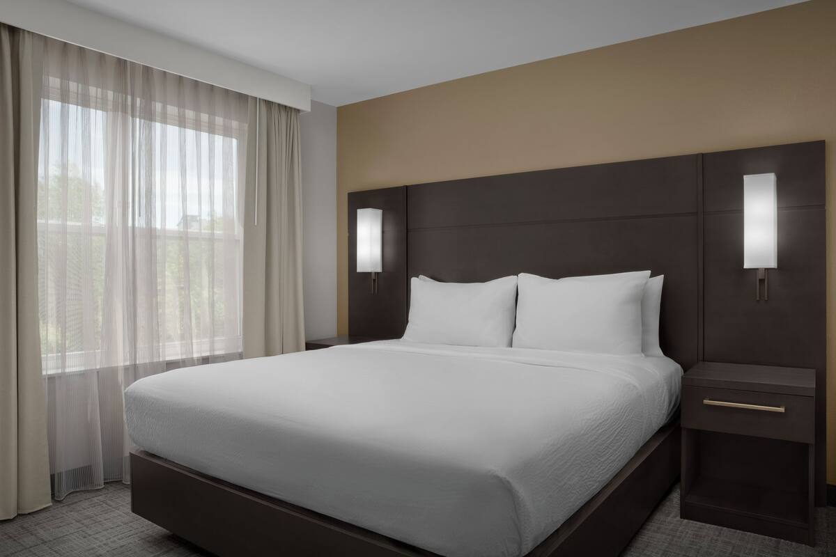 Een hotelkamer met een groot bed, twee nachtkastjes, een hoofdeinde met ingebouwde verlichting en dunne gordijnen voor het raam.