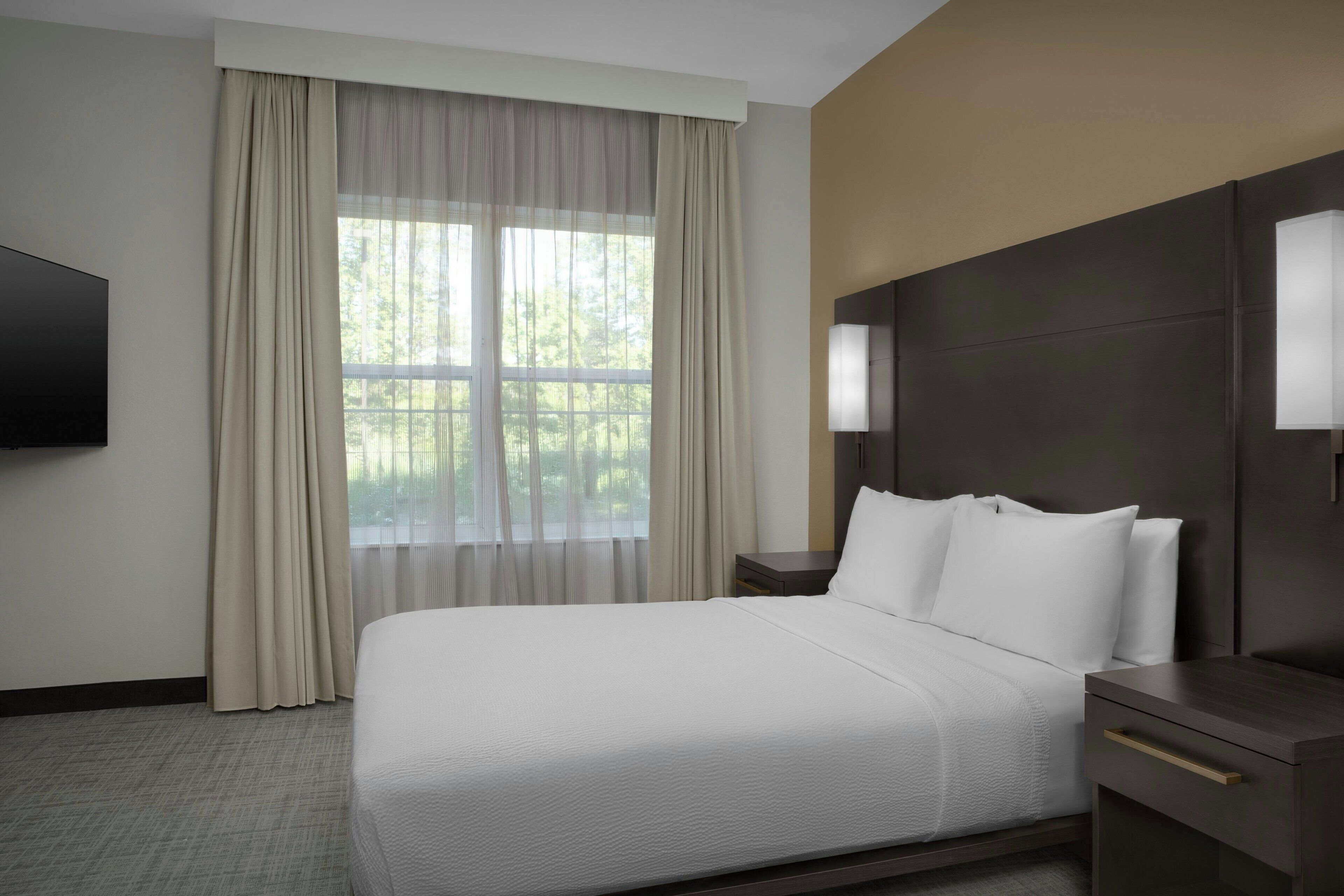 Quarto de hotel com cama grande, televisão, mesa de cabeceira e janela com cortinas.