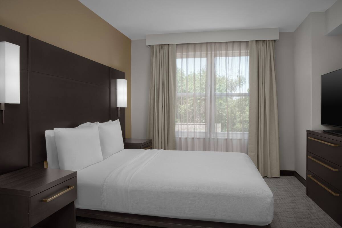 Een hotelkamer met een groot bed, een televisie, een nachtkastje en een raam met gordijnen.