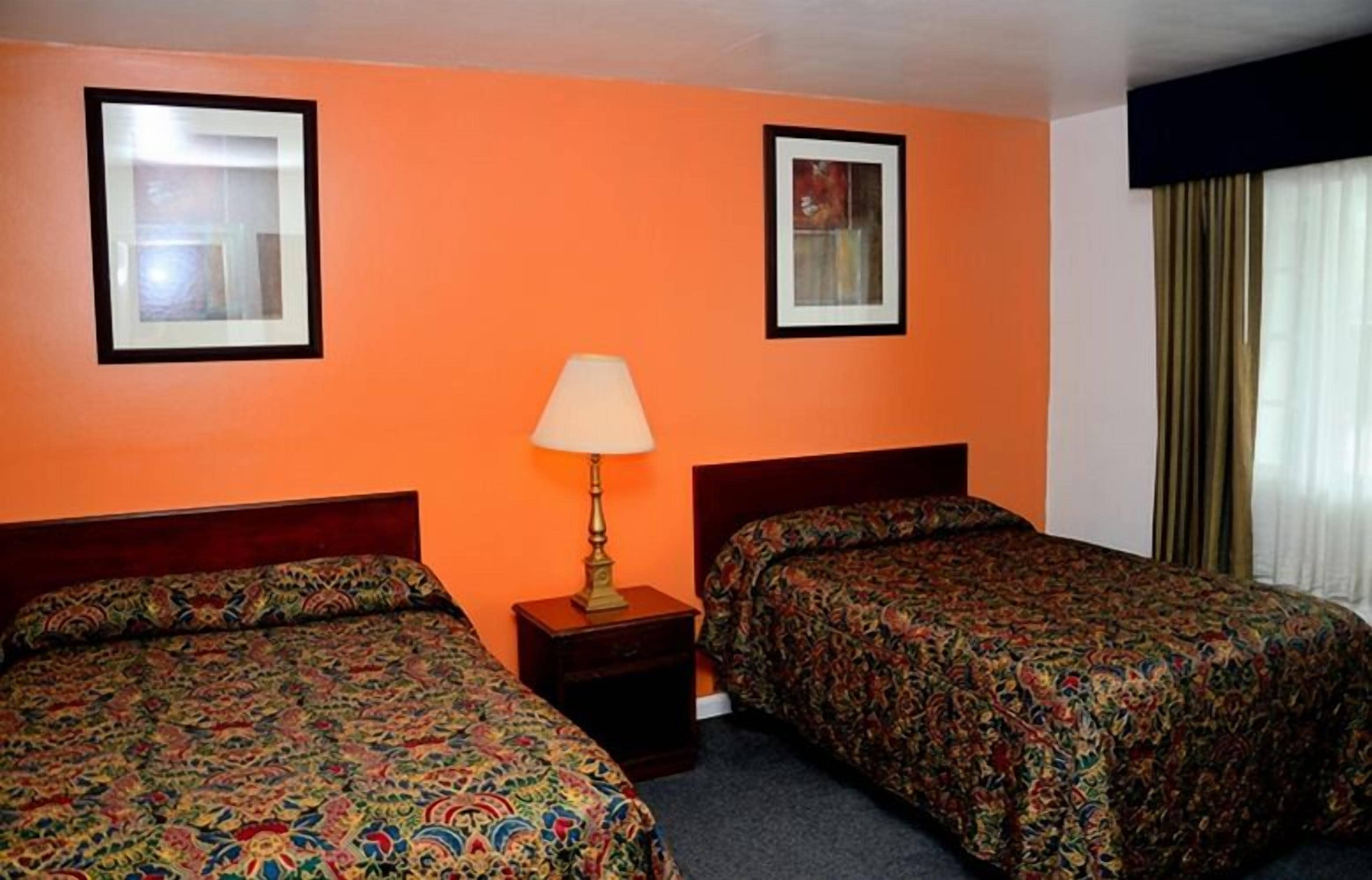 Une chambre d’hôtel avec deux lits, une table de chevet avec une lampe et deux tableaux encadrés sur les murs orange.