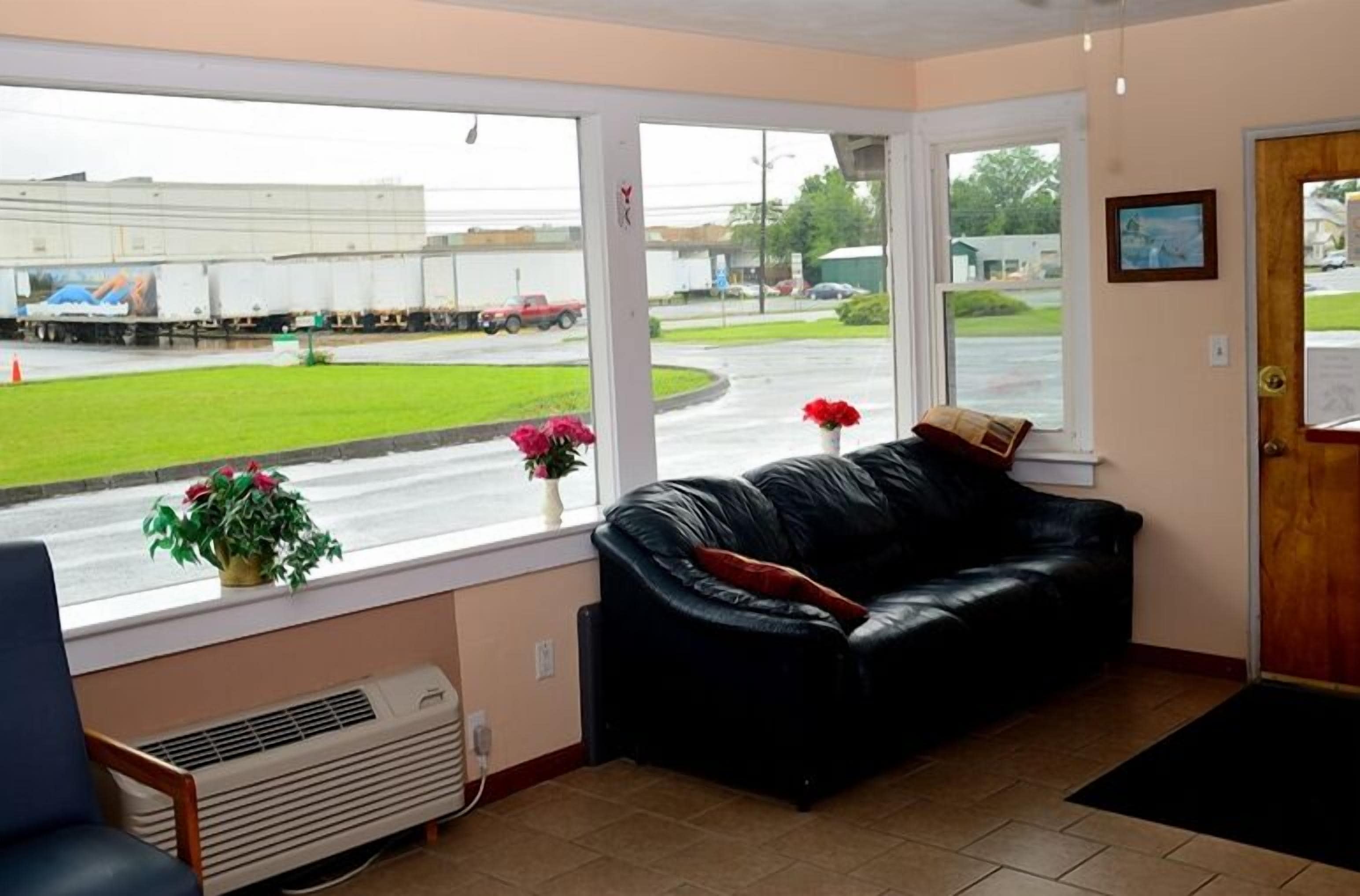 Una habitación con un sofá de cuero negro, una ventana que da a una calle con camiones, y una puerta con un cuadro enmarcado.
