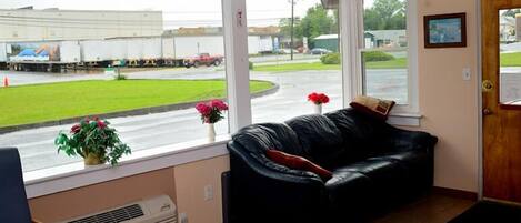A room with a black leather sofa, a window with a view of a road and trucks, and a door with a framed picture.
